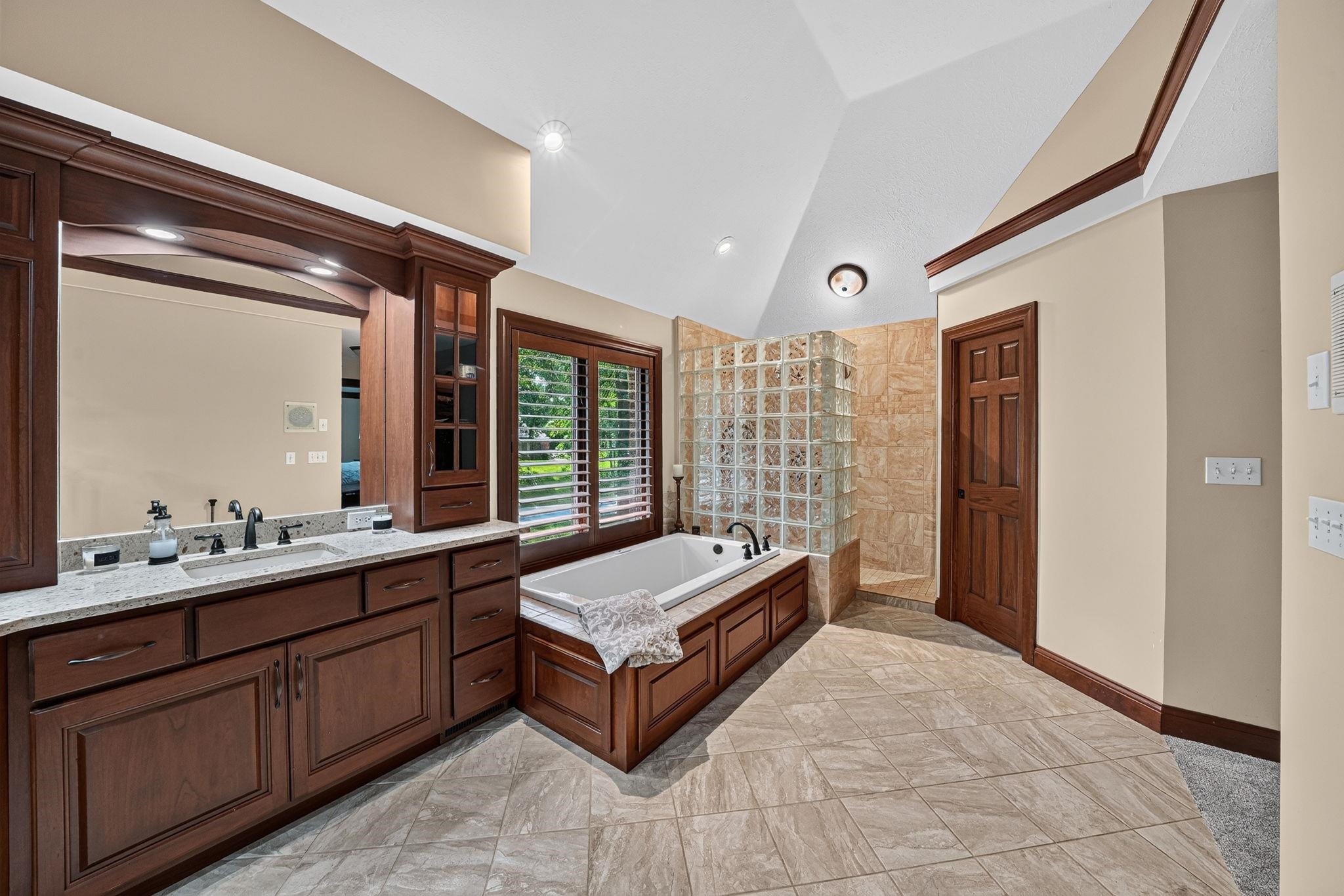 Spacious bathroom featuring a double vanity with stone countertop, large soaking tub, glass block shower enclosure, and tiled flooring