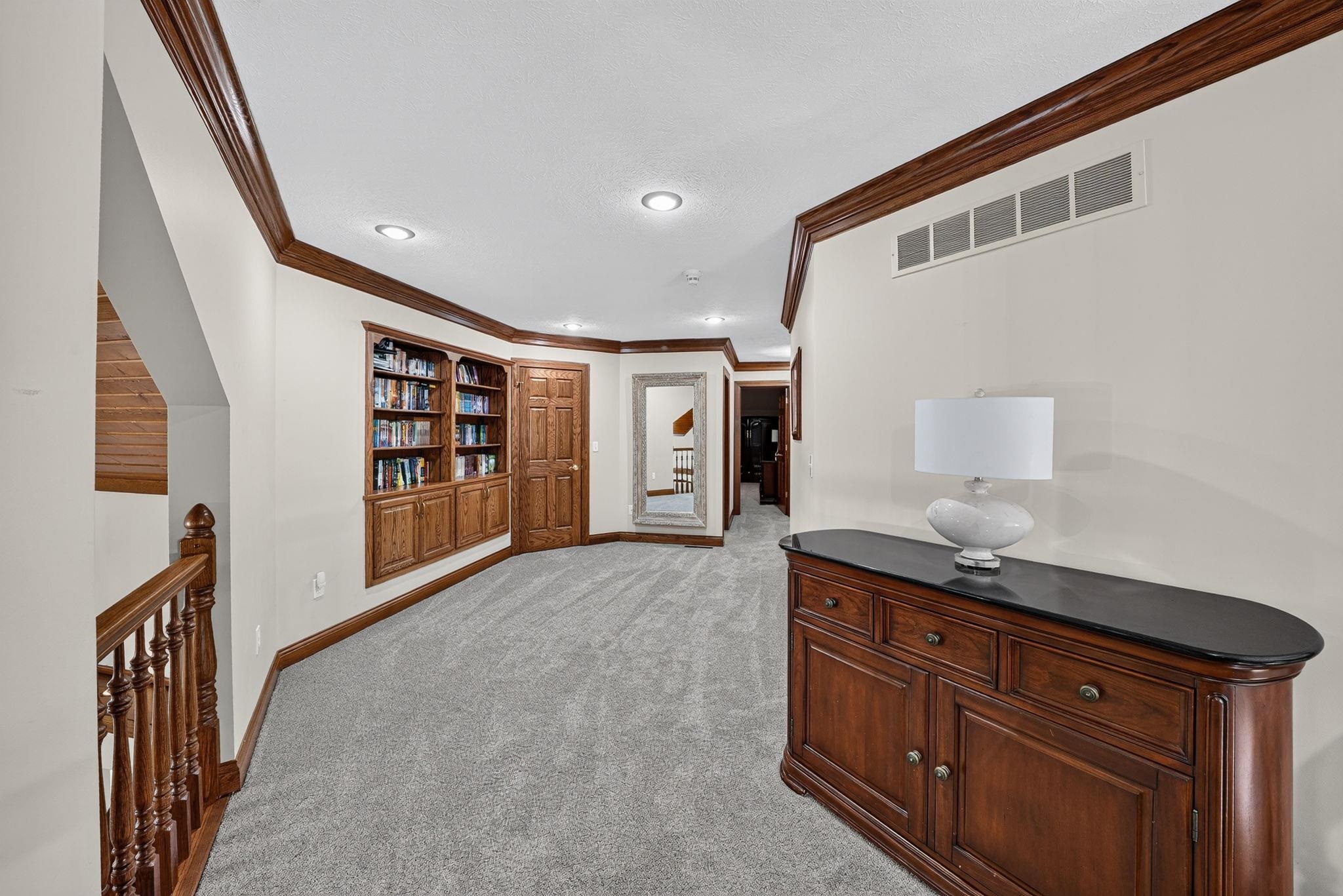 Hallway featuring light gray carpeting, recessed ceiling lighting, built-in wood cabinetry with shelving, a wood-finish staircase railing, and dark wood trim