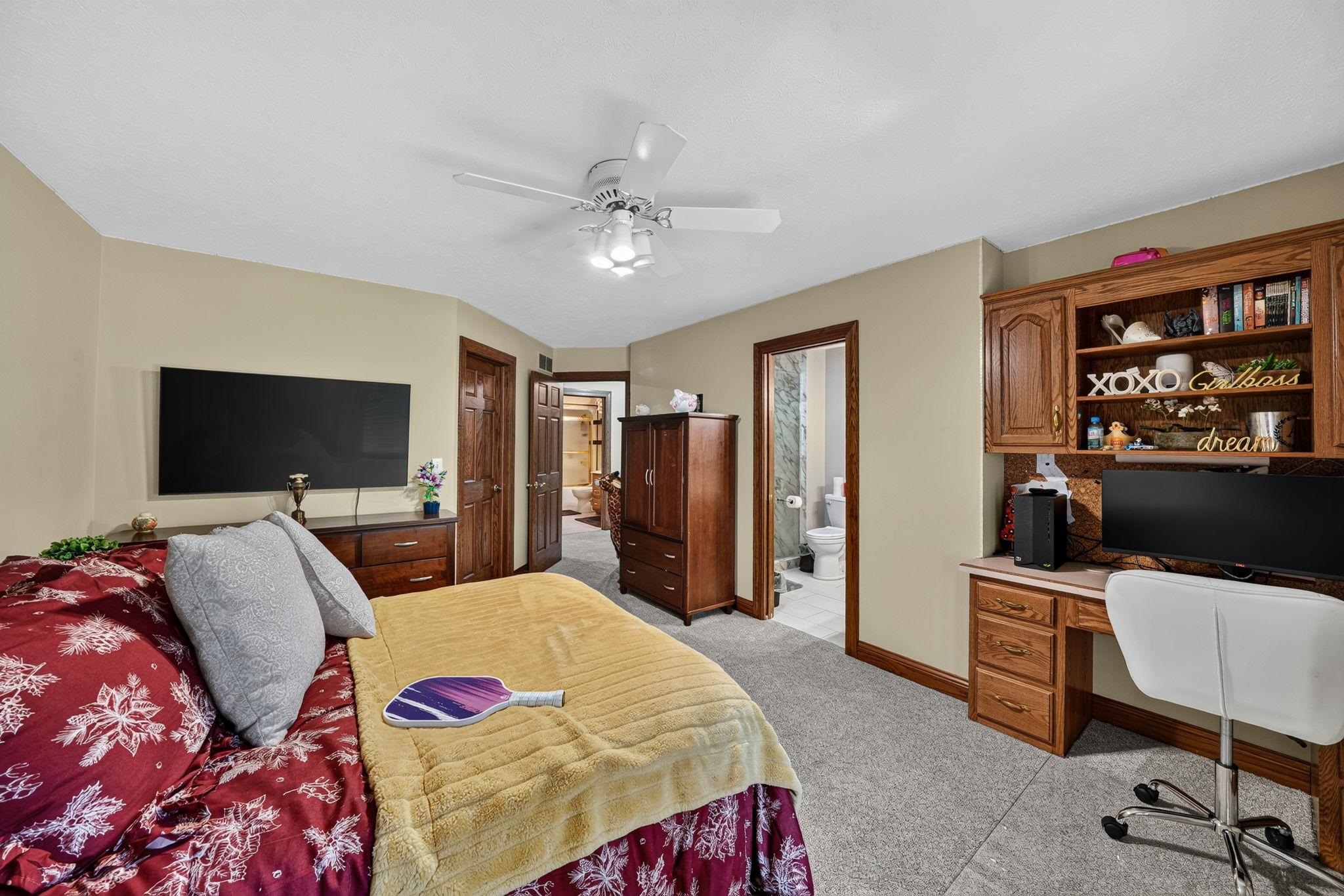 Carpeted bedroom featuring a built-in desk with overhead shelving, a ceiling fan with integrated lighting, wood-finish trim, and a wall-mounted flat-screen television
