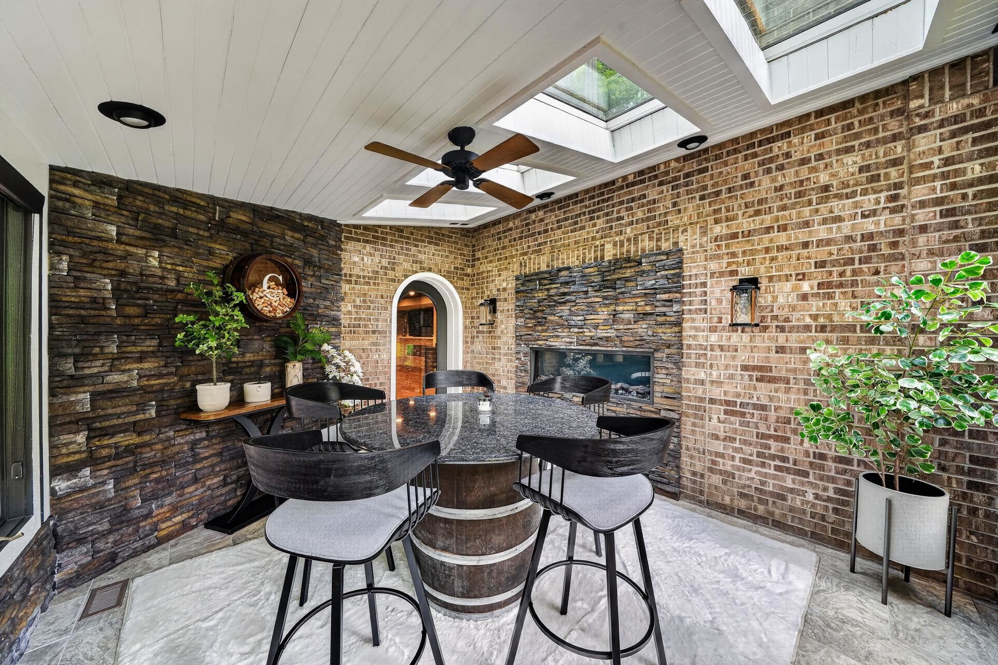 Covered patio featuring a stone accent wall, brick walls, an integrated fireplace, skylights, and a wood-slat ceiling with a ceiling fan