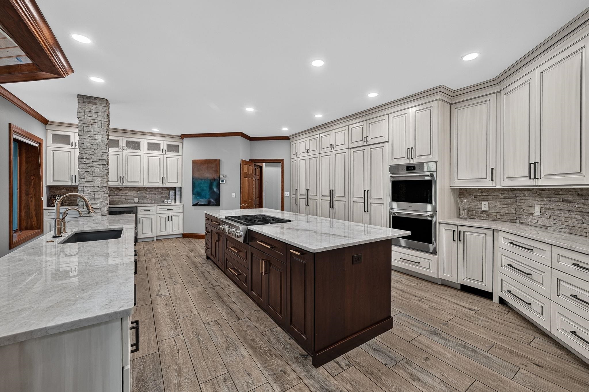 Gourmet kitchen featuring a large center island with a gas range, dual wall ovens, extensive light-toned cabinetry, stone countertops, and wood-finish flooring