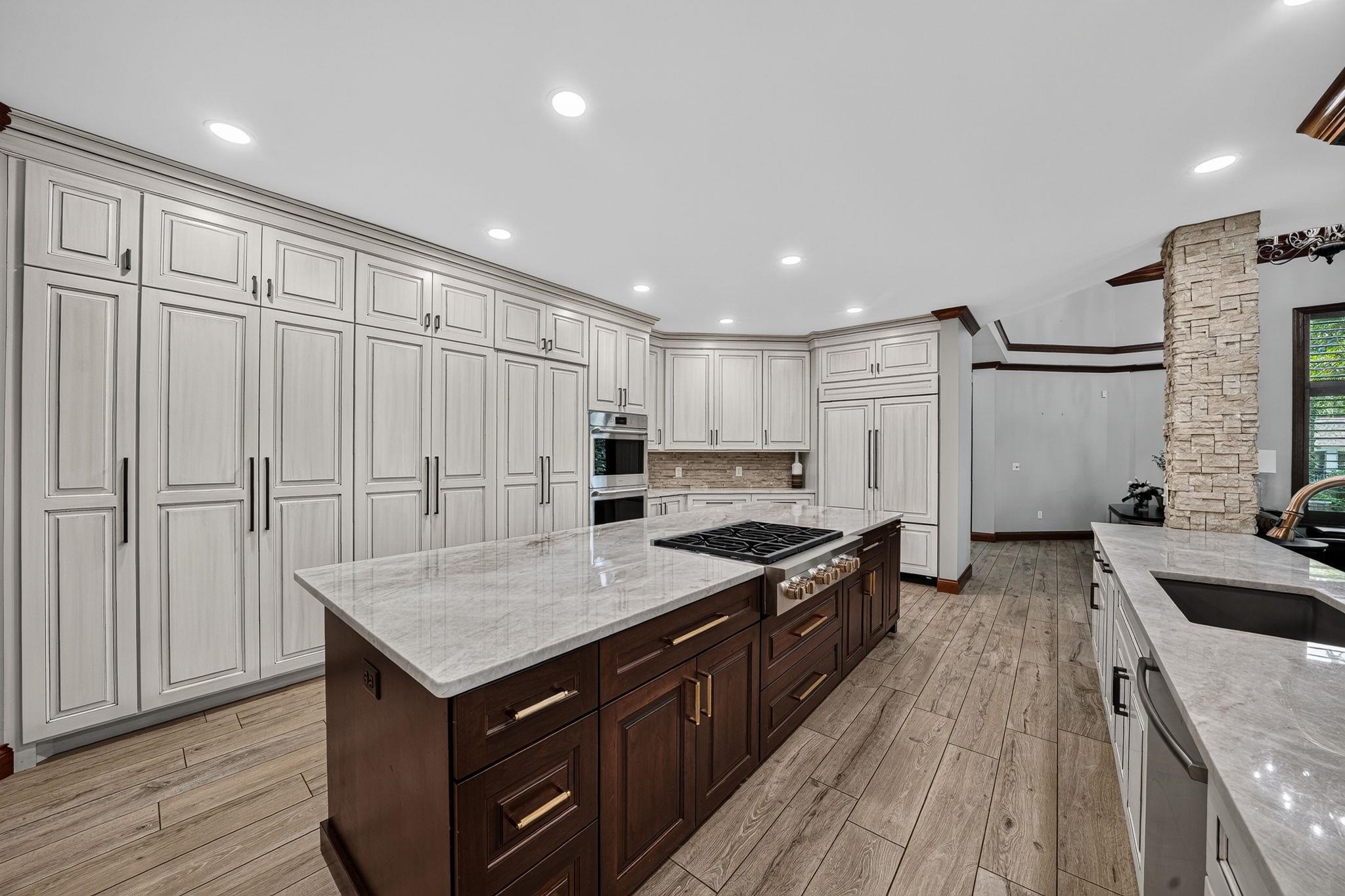 Spacious kitchen featuring a large central island with a gas cooktop, extensive light-toned cabinetry, built-in appliances, wood-finish flooring, and stone accent walls