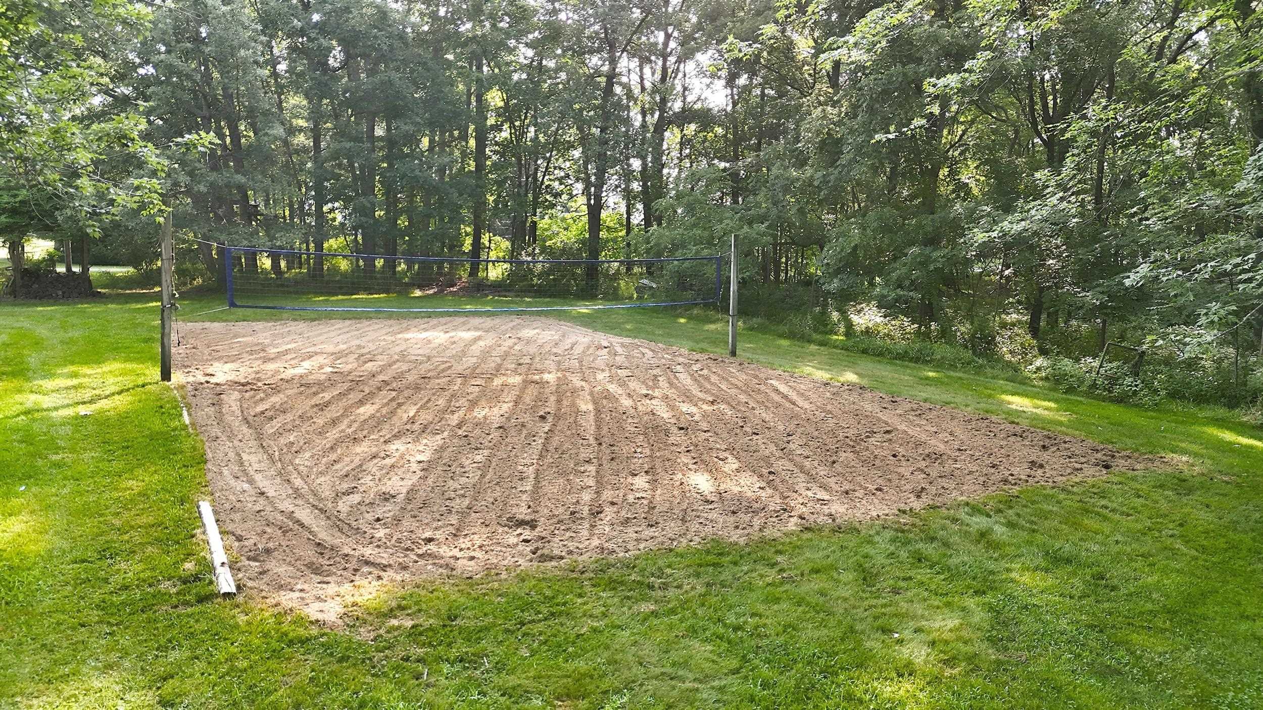 Sand volleyball court set within a grassy area, bordered by mature trees