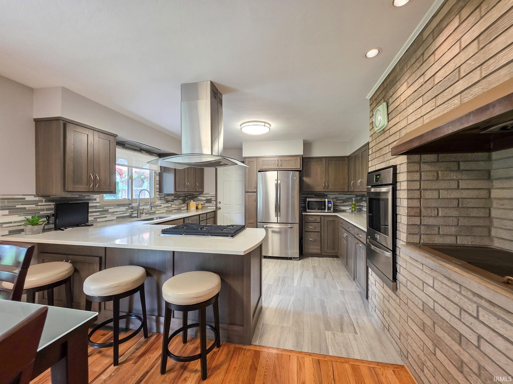 Spacious kitchen featuring a built-in gas cooktop and stainless steel range hood, ample cabinetry, stainless steel appliances, and a brick accent wall with an integrated oven
