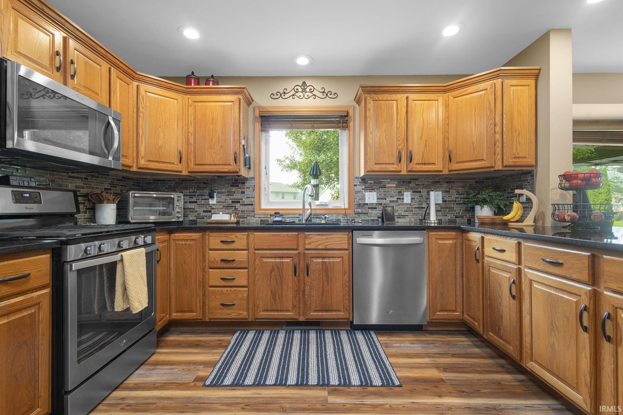 Kitchen featuring wood cabinetry, dark countertops, mosaic tile backsplash, stainless steel appliances, and wood-finish flooring