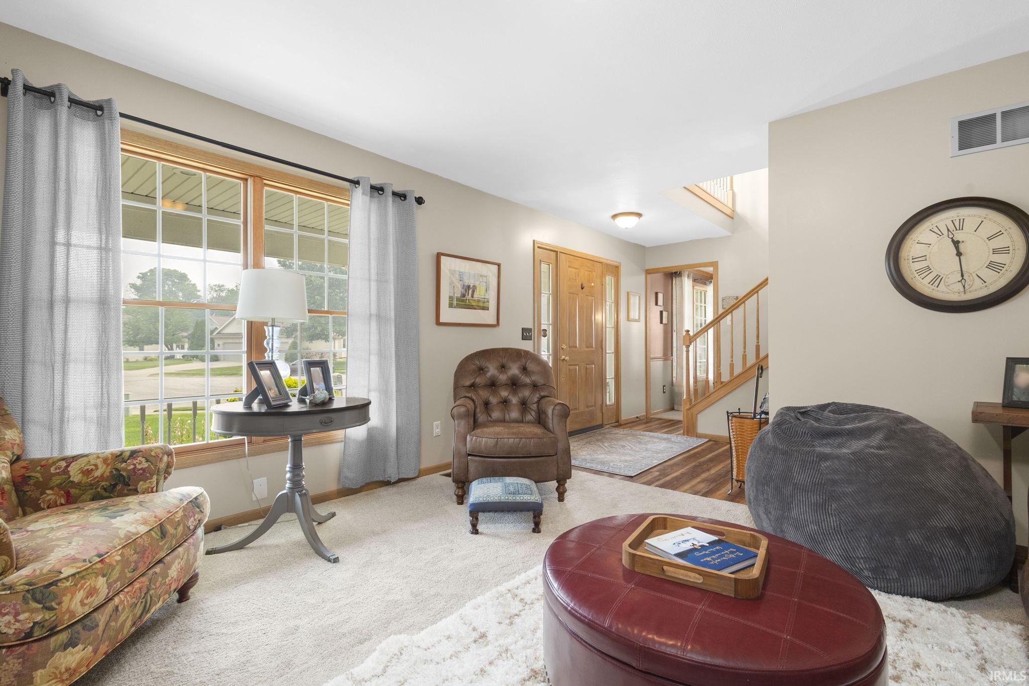 Bright living area featuring light-colored carpet, large window with grid pattern, and a natural wood-finish front door