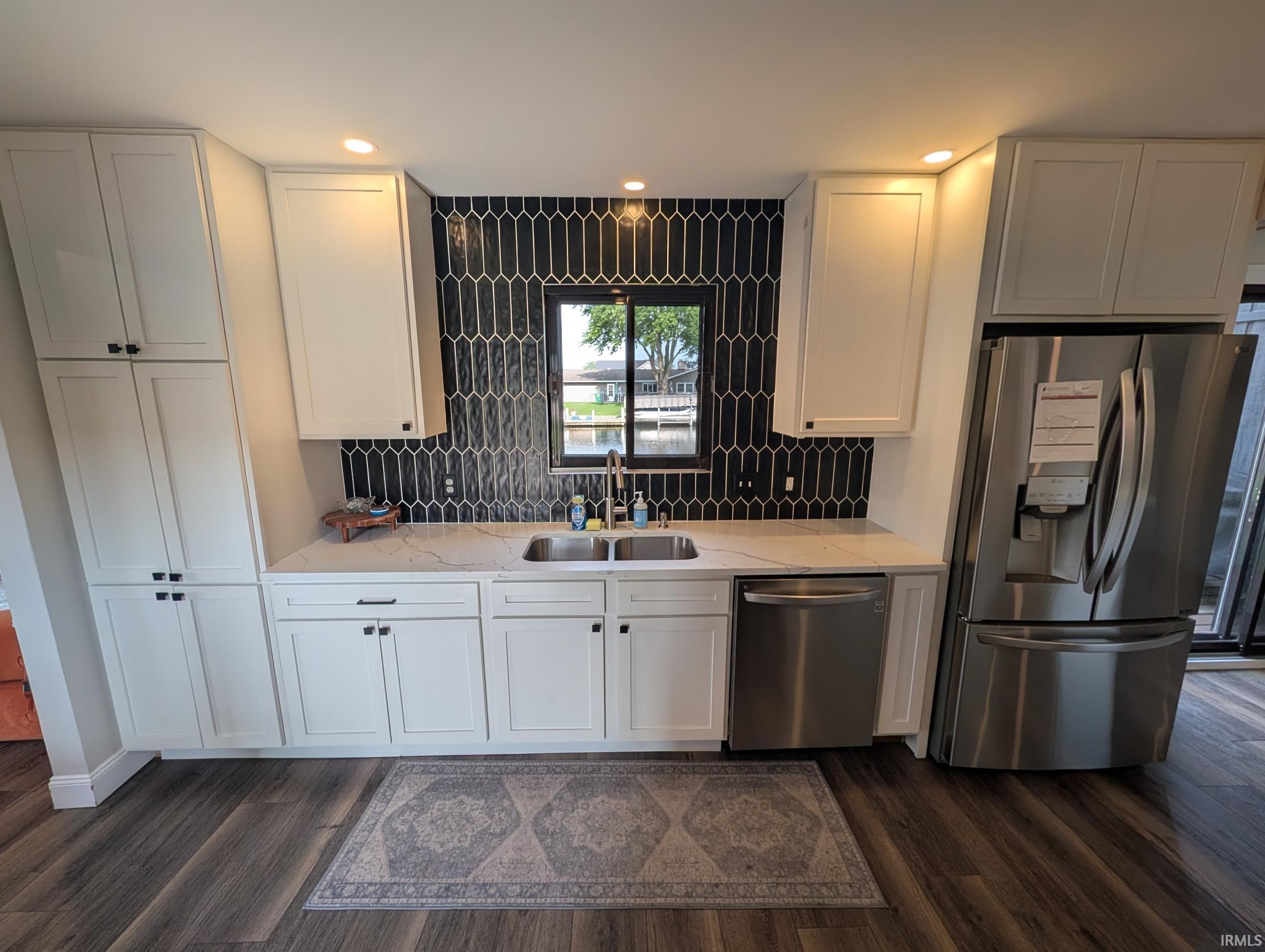 Kitchen featuring white cabinetry, a black elongated hexagonal tile backsplash, a stainless steel double basin sink, light-toned countertops, and wood-finish flooring