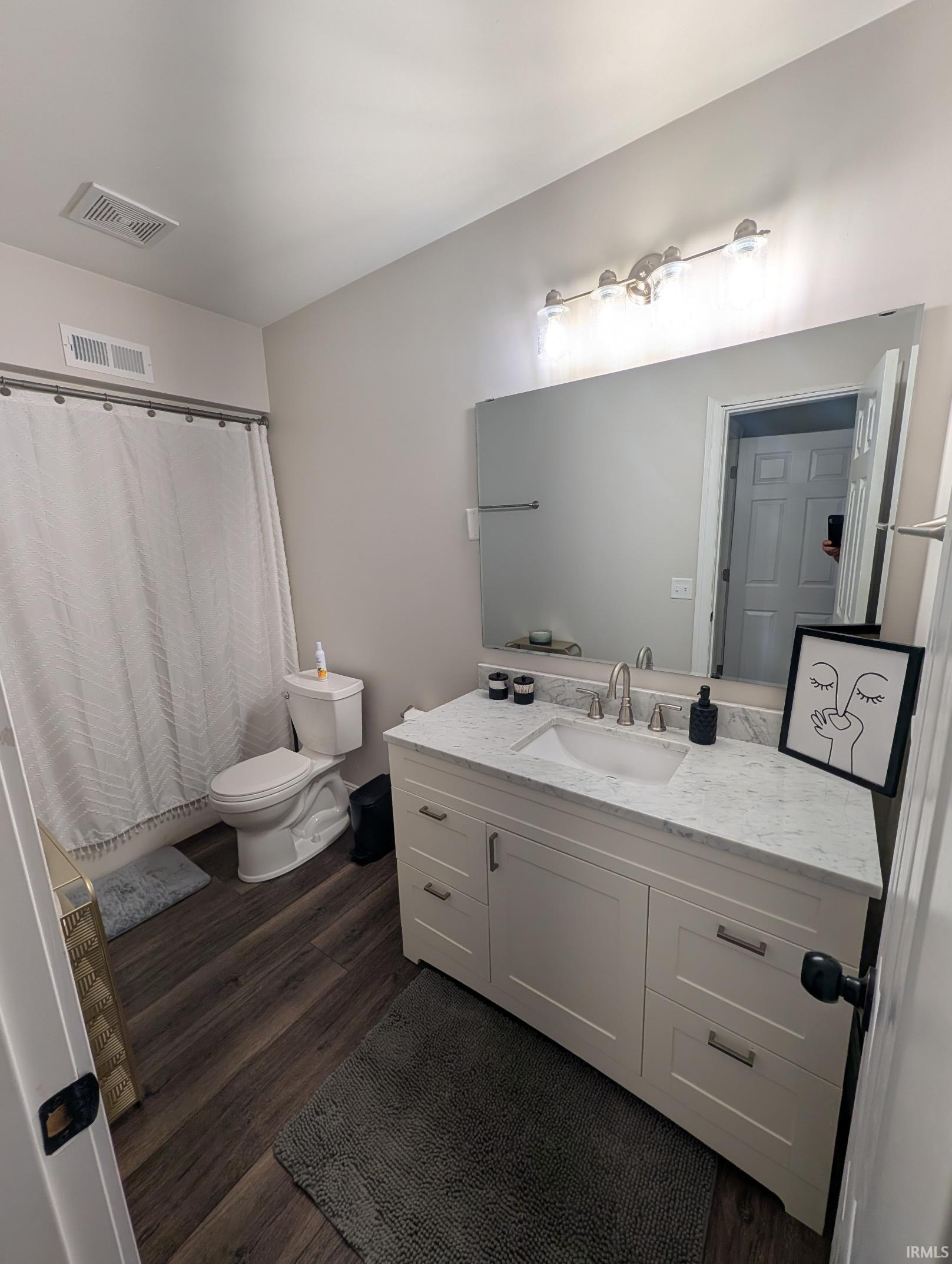 Bathroom featuring a single-sink vanity with a light-toned countertop, wood-finish flooring, a toilet, a shower/tub combination with a curtain rod, and a large wall mirror
