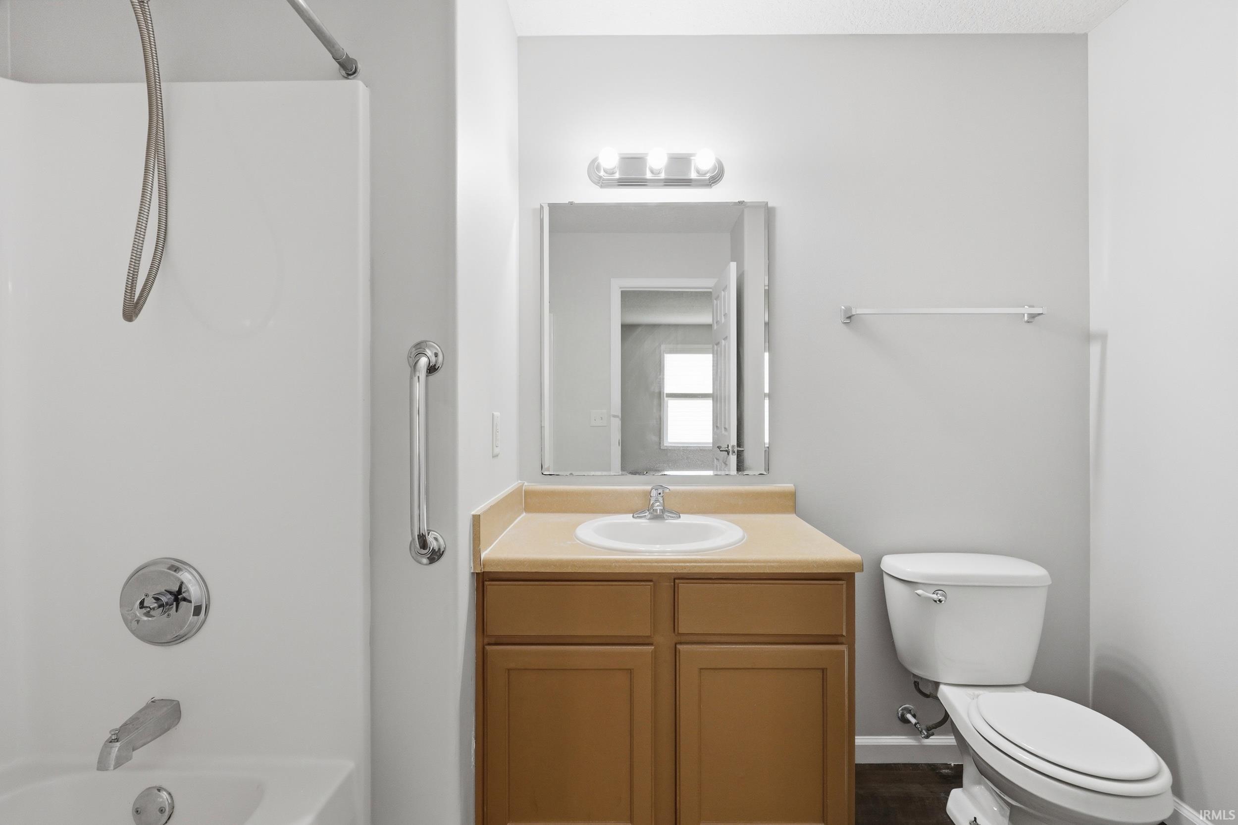 Full bathroom featuring a white tub/shower combination with grab bar, a single vanity with wood-finish cabinetry and a light-colored countertop, and a white toilet