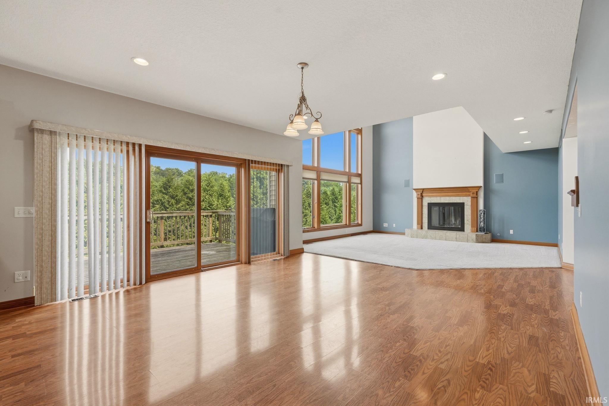 Expansive open-concept living area featuring rich wood-finish flooring, a two-story fireplace with a wood mantle, a wall of large windows, and a glass sliding door opening to an outdoor deck