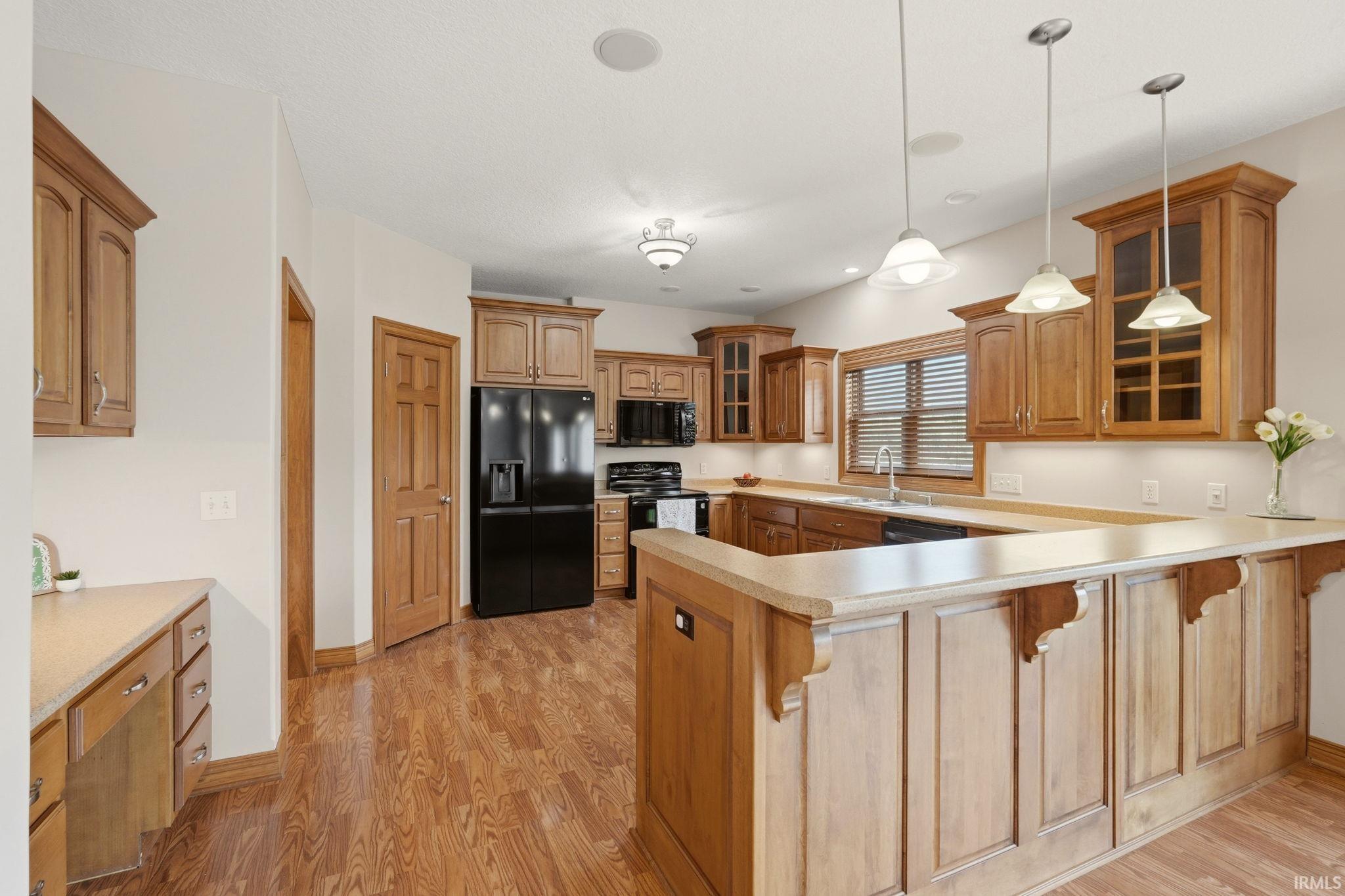 Kitchen featuring wood-finish flooring, light wood cabinetry, U-shaped countertops, integrated black appliances, and pendant lighting over a breakfast bar