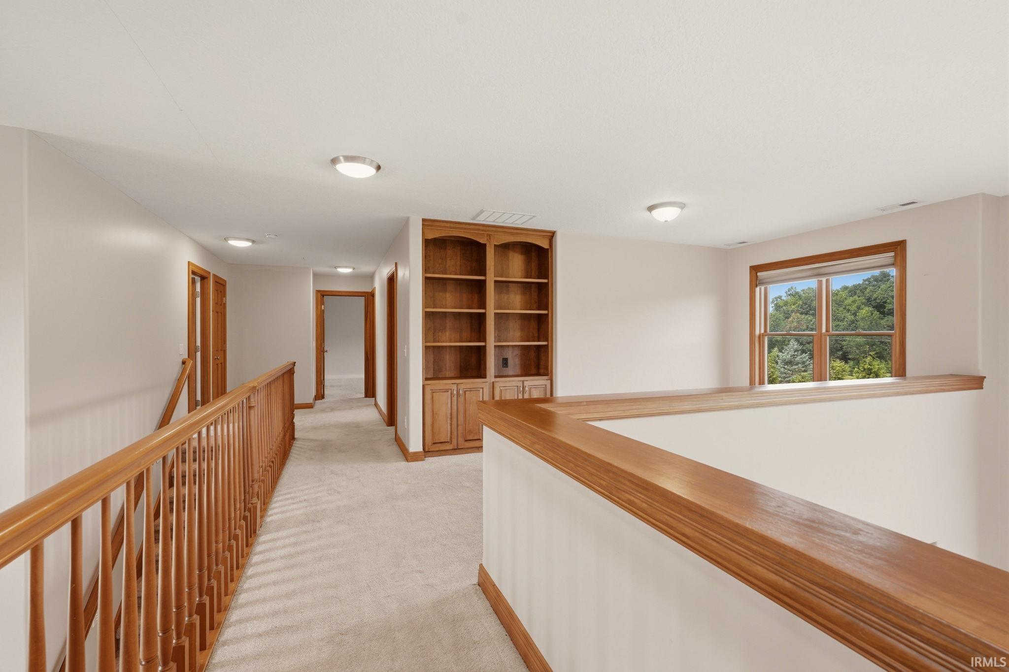 Carpeted hallway featuring a wood-toned handrail, built-in wooden shelving with lower cabinetry, and a window with wood trim