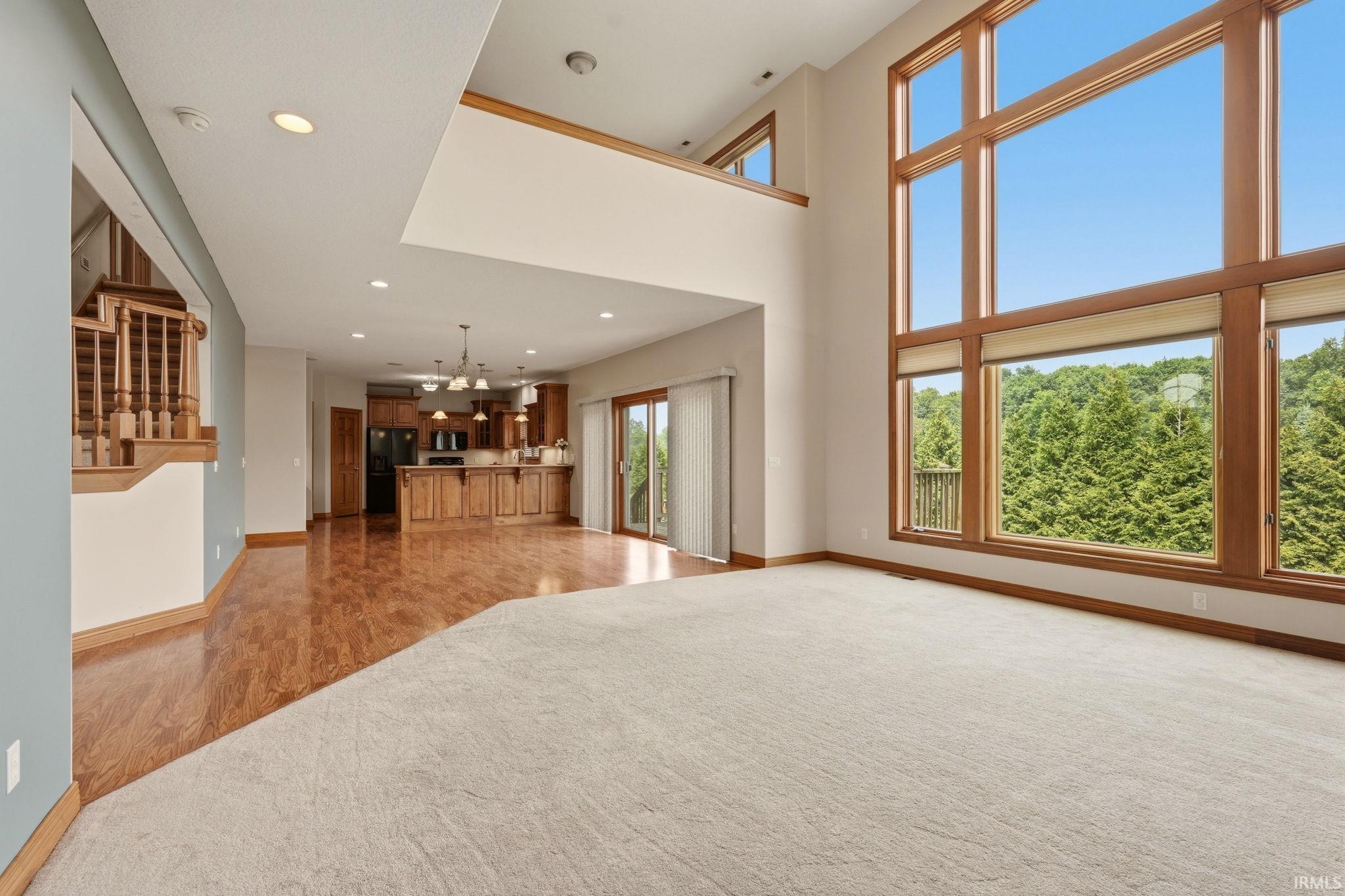 Open-concept living area featuring a two-story ceiling, large picture windows, wood-finish flooring, light-toned carpeting, and a visible kitchen with wood cabinetry