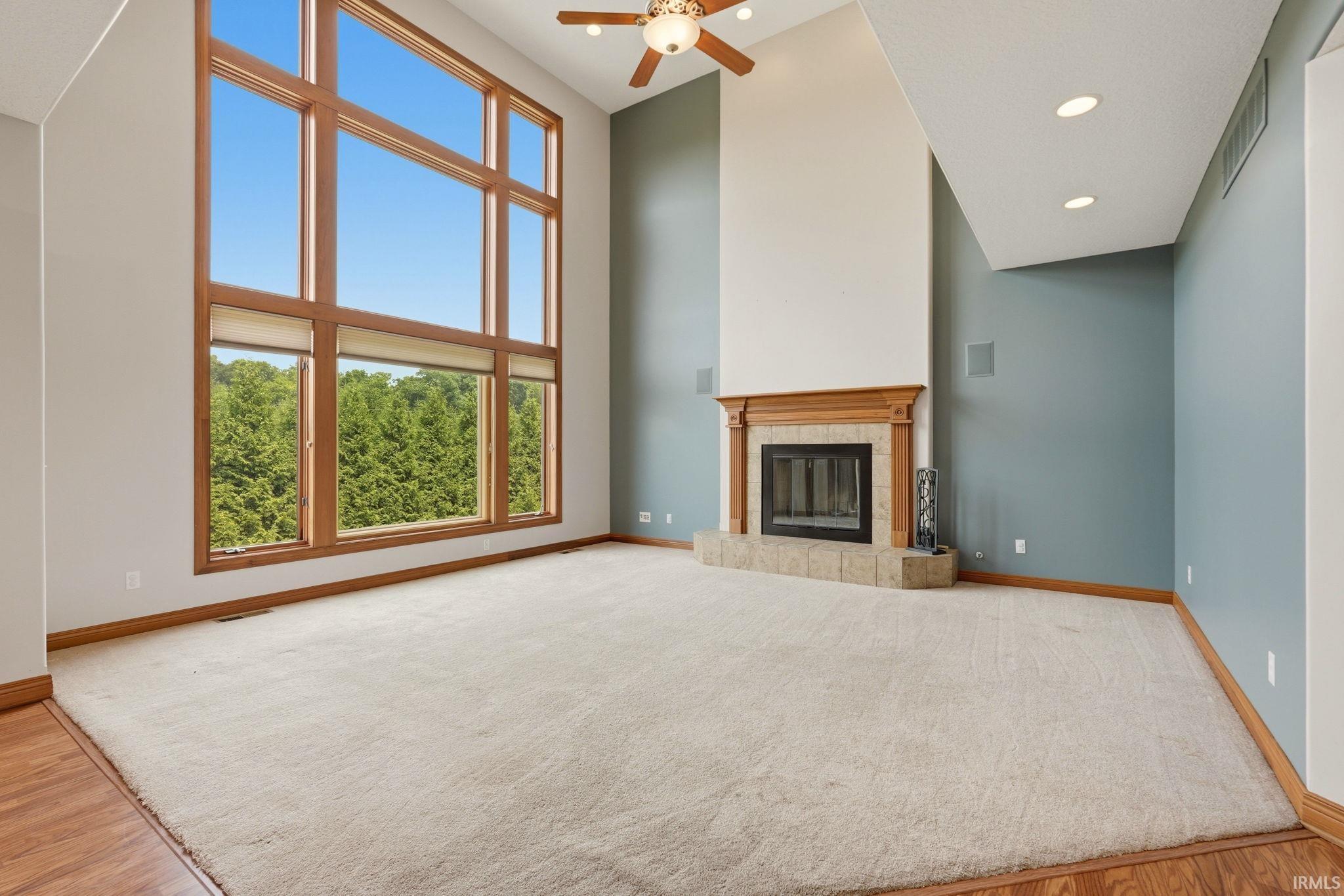 Two-story great room featuring a tall window wall, wood-finish flooring, a ceiling fan, and a fireplace with a wood mantel and stone surround