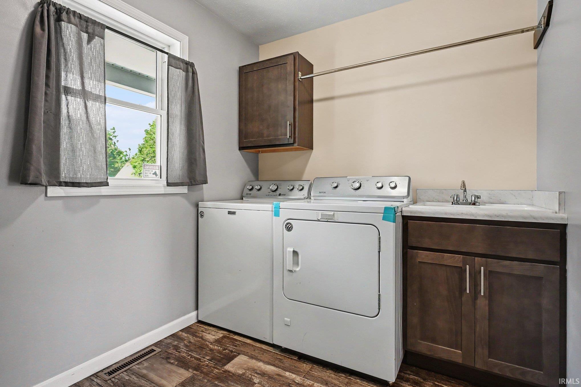Dedicated laundry area featuring wood-finish flooring, a white trim window, a dark wood-finish wall cabinet, a stainless steel hanging rod, and a utility sink with a stone-finish countertop and dark wood-finish base cabinetry