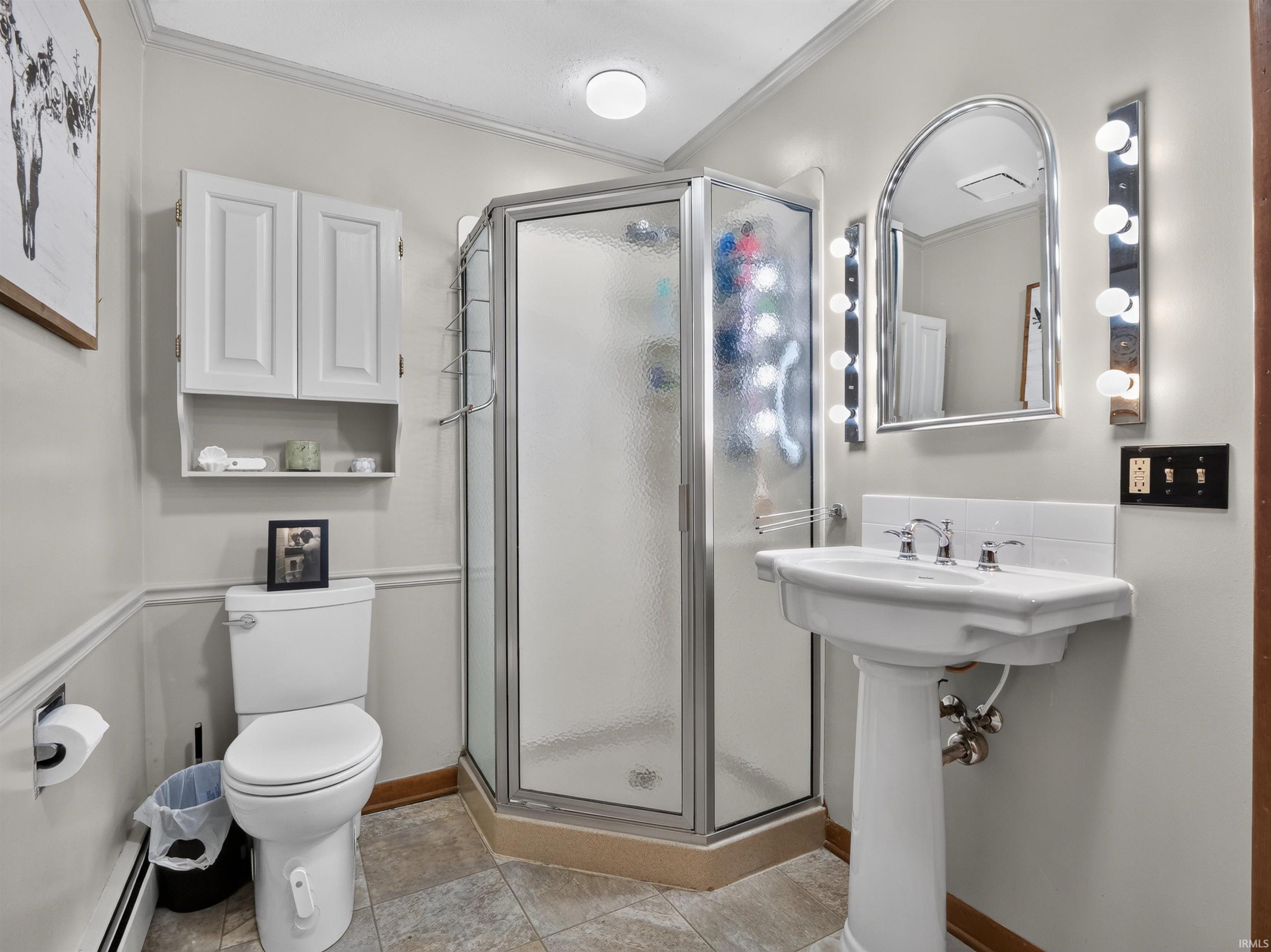 Pedestal sink with chrome fixtures, arched mirror with vanity lighting, and corner glass shower enclosure