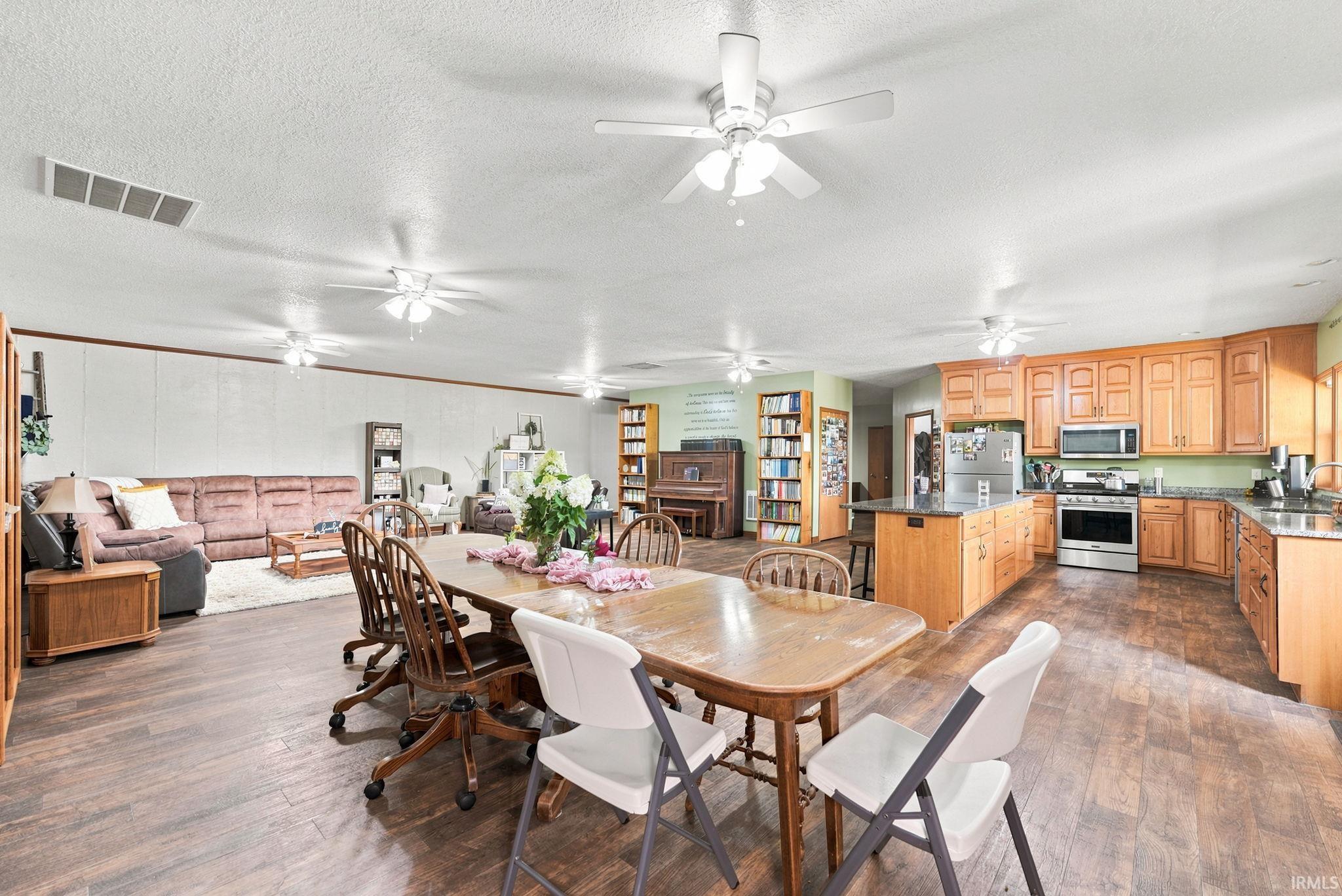 Spacious open-concept living area featuring wood-finish flooring, a kitchen with wood cabinetry and stainless steel appliances, a center island with bar seating, and multiple ceiling fans