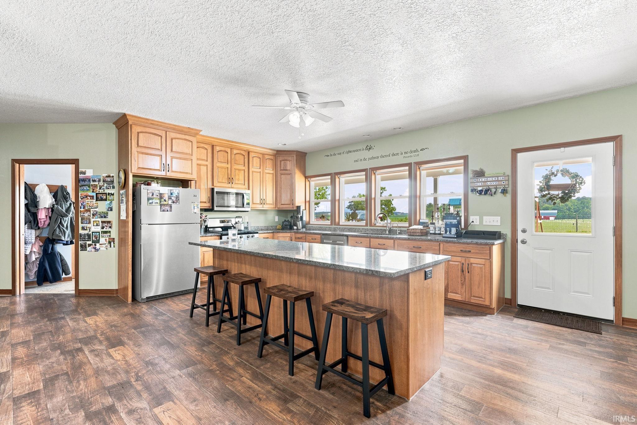 Open-concept kitchen featuring wood-finish flooring, light wood cabinetry, stainless steel appliances, and a central island with an extended countertop