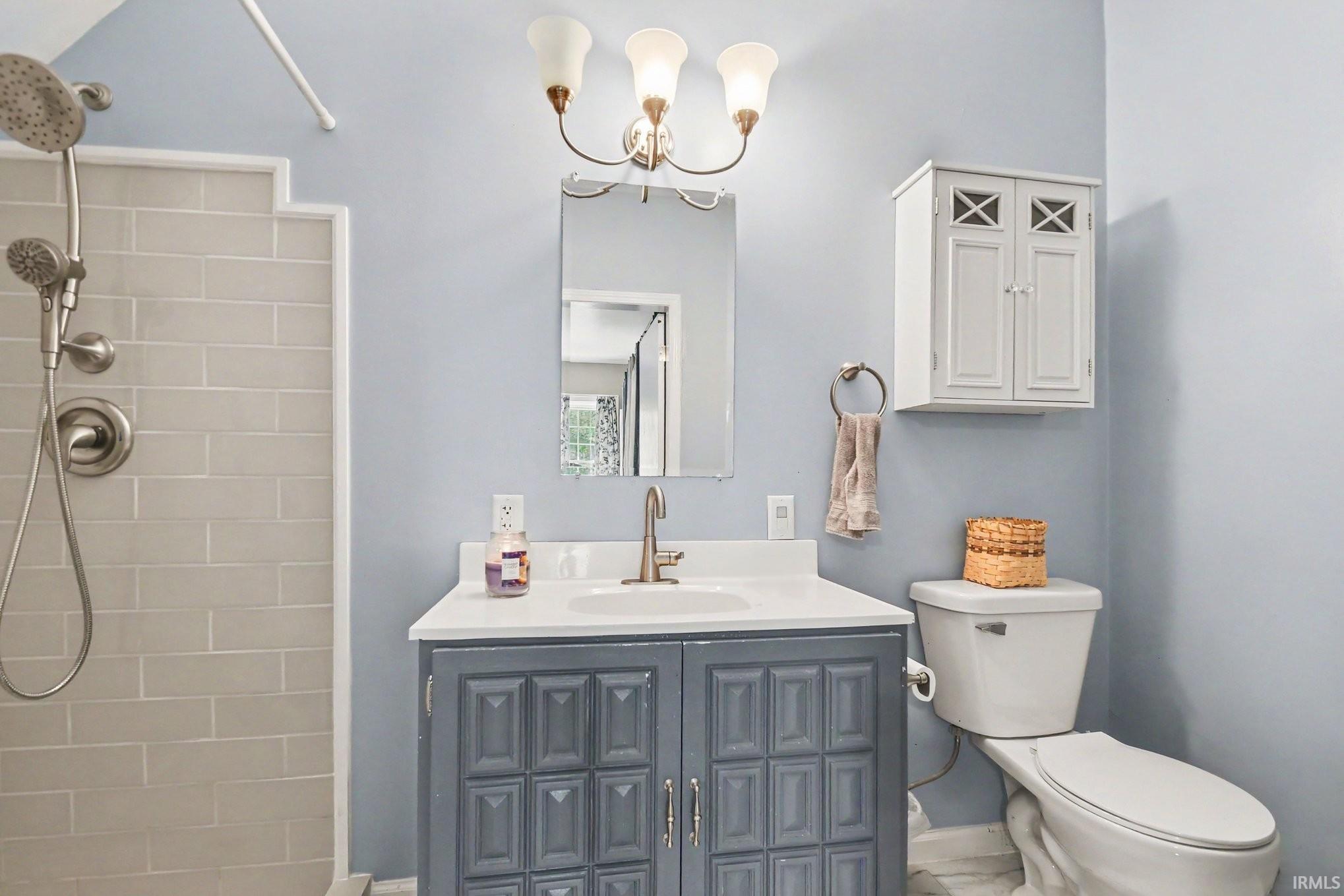 Bathroom featuring a tiled shower with brushed nickel fixtures, a vanity with a white countertop and dark gray cabinetry, a wall-mounted medicine cabinet, and a toilet