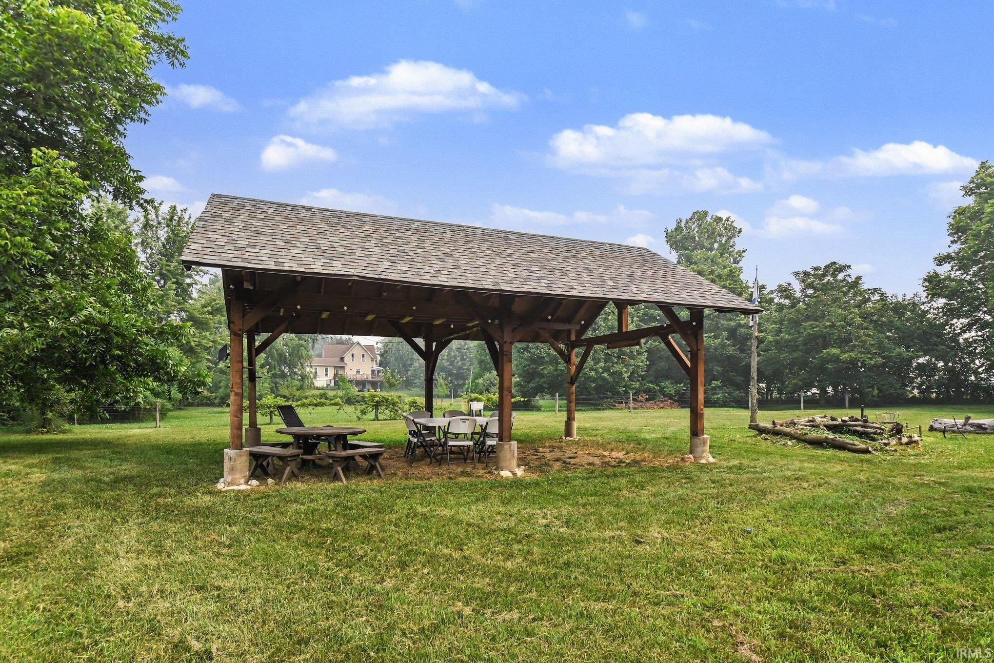 Outdoor wood-framed pavilion with a shingled roof, set on a grass lawn, featuring concrete pillar bases and a visible fire pit area