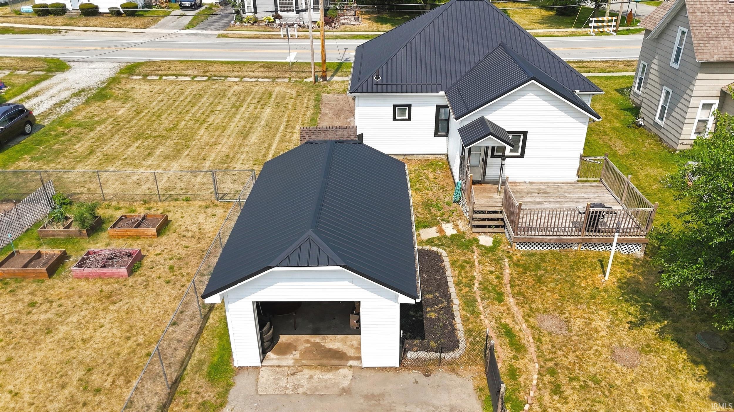 White siding residence featuring a dark metal roof, detached garage with matching metal roof, rear wooden deck, and a fenced yard with raised garden beds