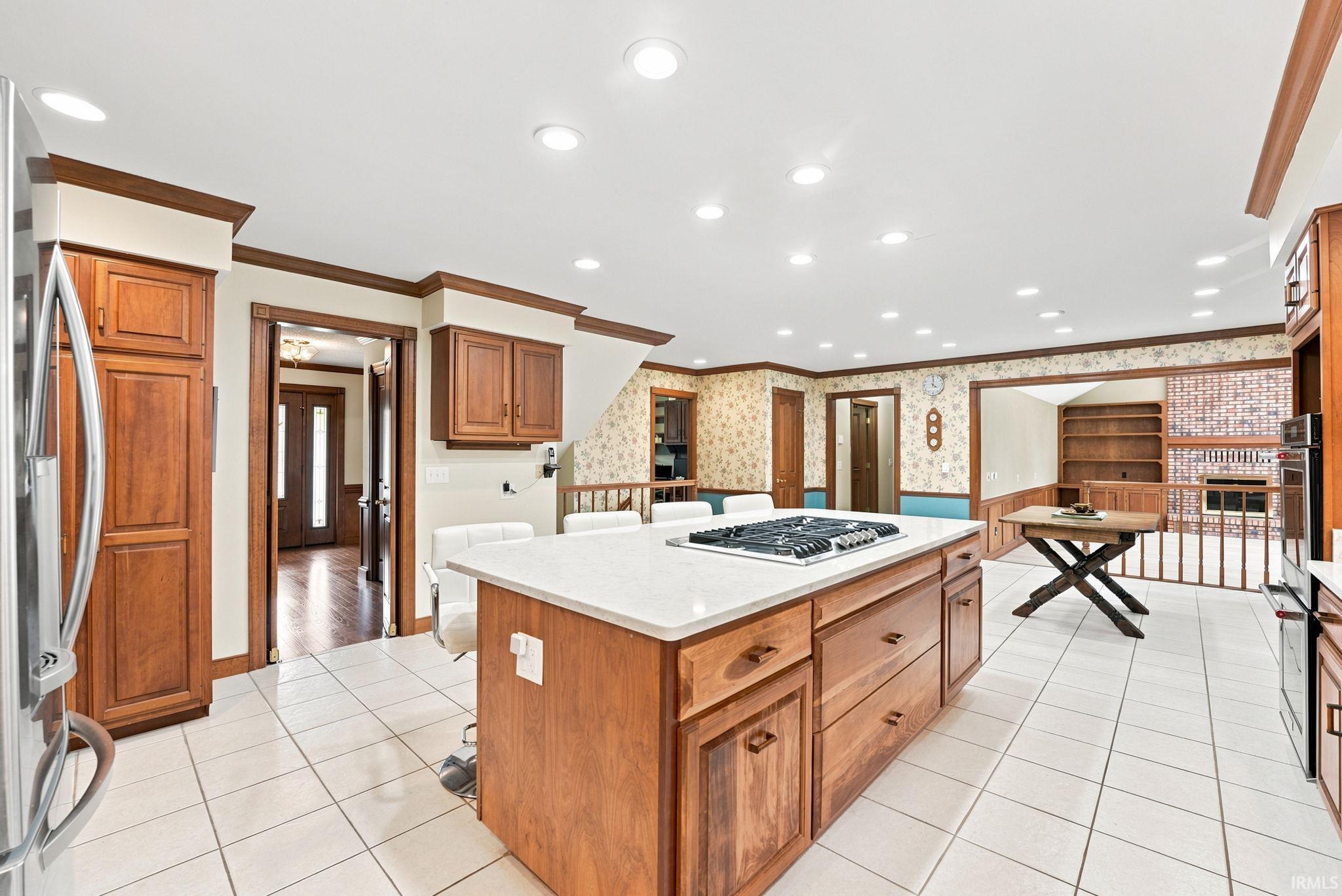 Spacious kitchen featuring a central island with an integrated cooktop and solid surface countertop, warm wood cabinetry, a stainless steel refrigerator, recessed lighting, and white tile flooring