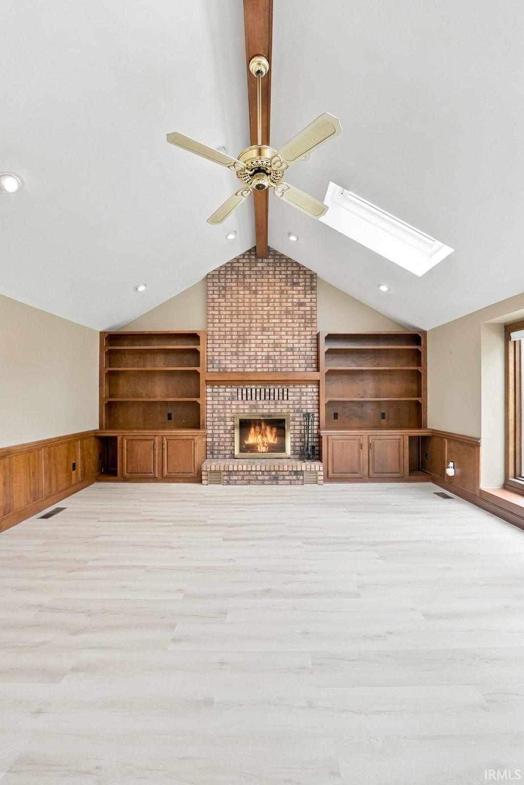 Vaulted ceiling living space featuring a brick fireplace, light wood-finish flooring, built-in shelving, and wood wainscoting