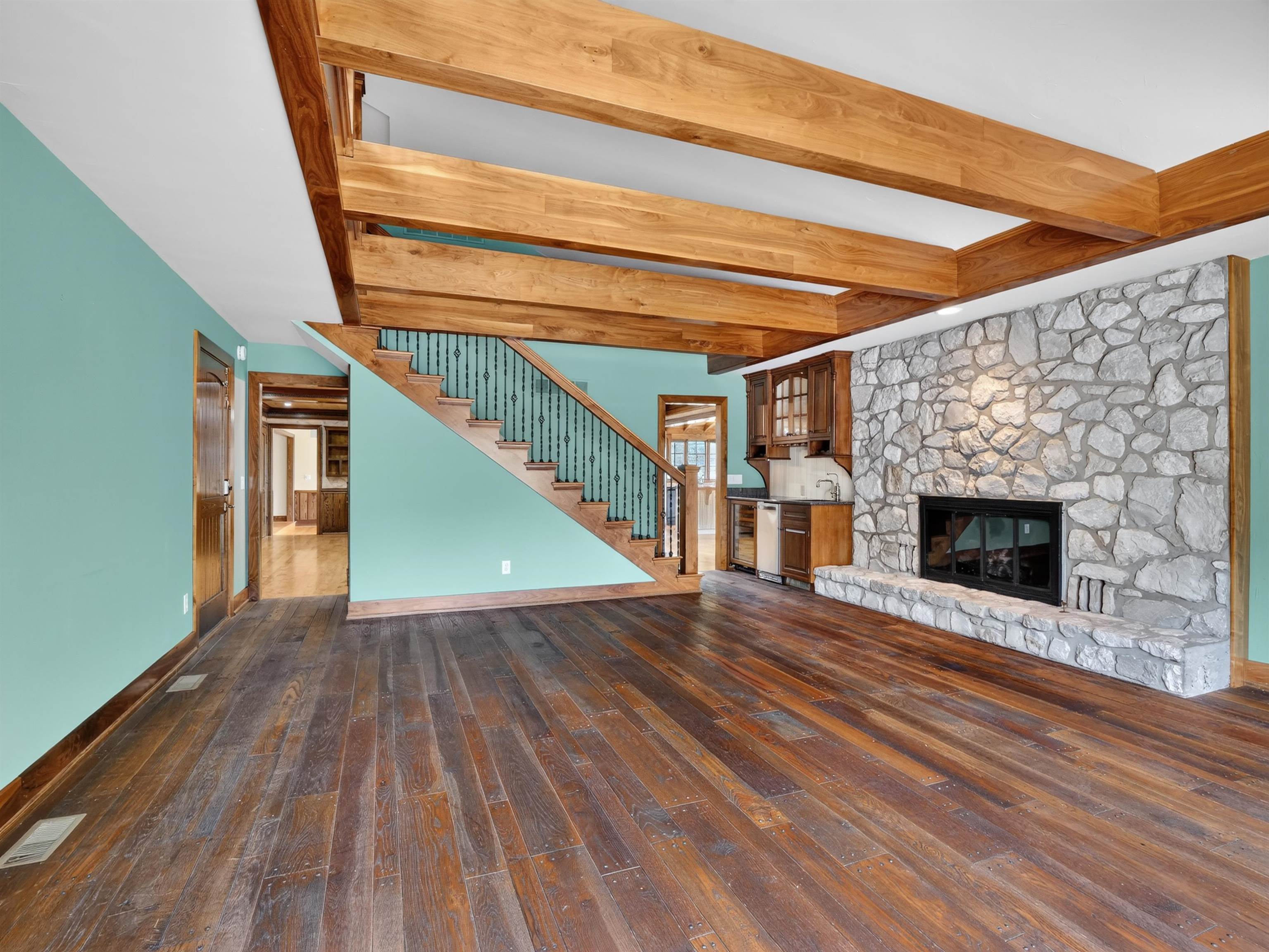Spacious interior featuring a stone fireplace, wood-finish flooring, exposed ceiling beams, a built-in wet bar, and a staircase with metal balusters