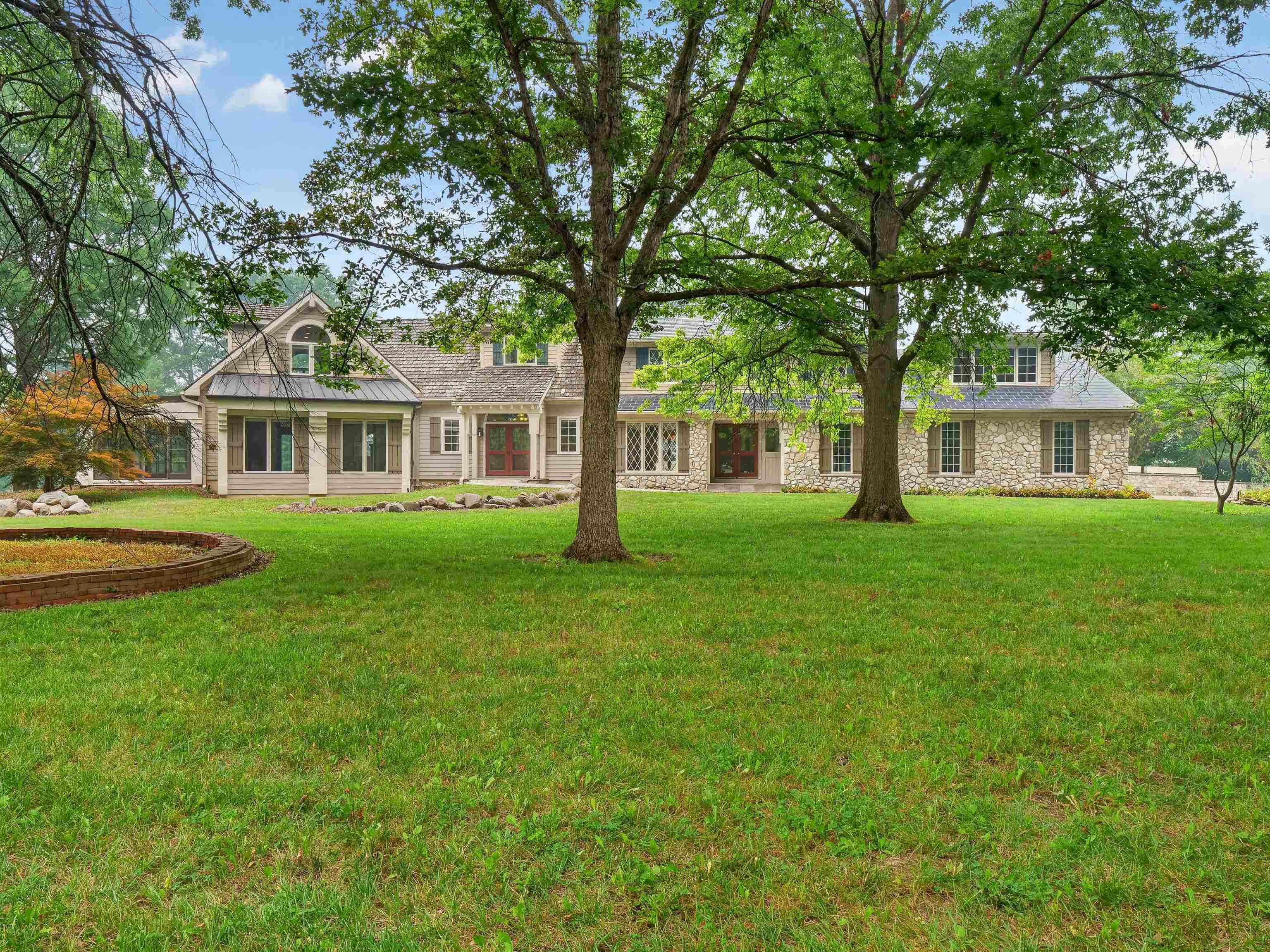 Spacious lawn with mature trees, stone and siding exterior, multiple dormer windows, shuttered windows, and double entry doors