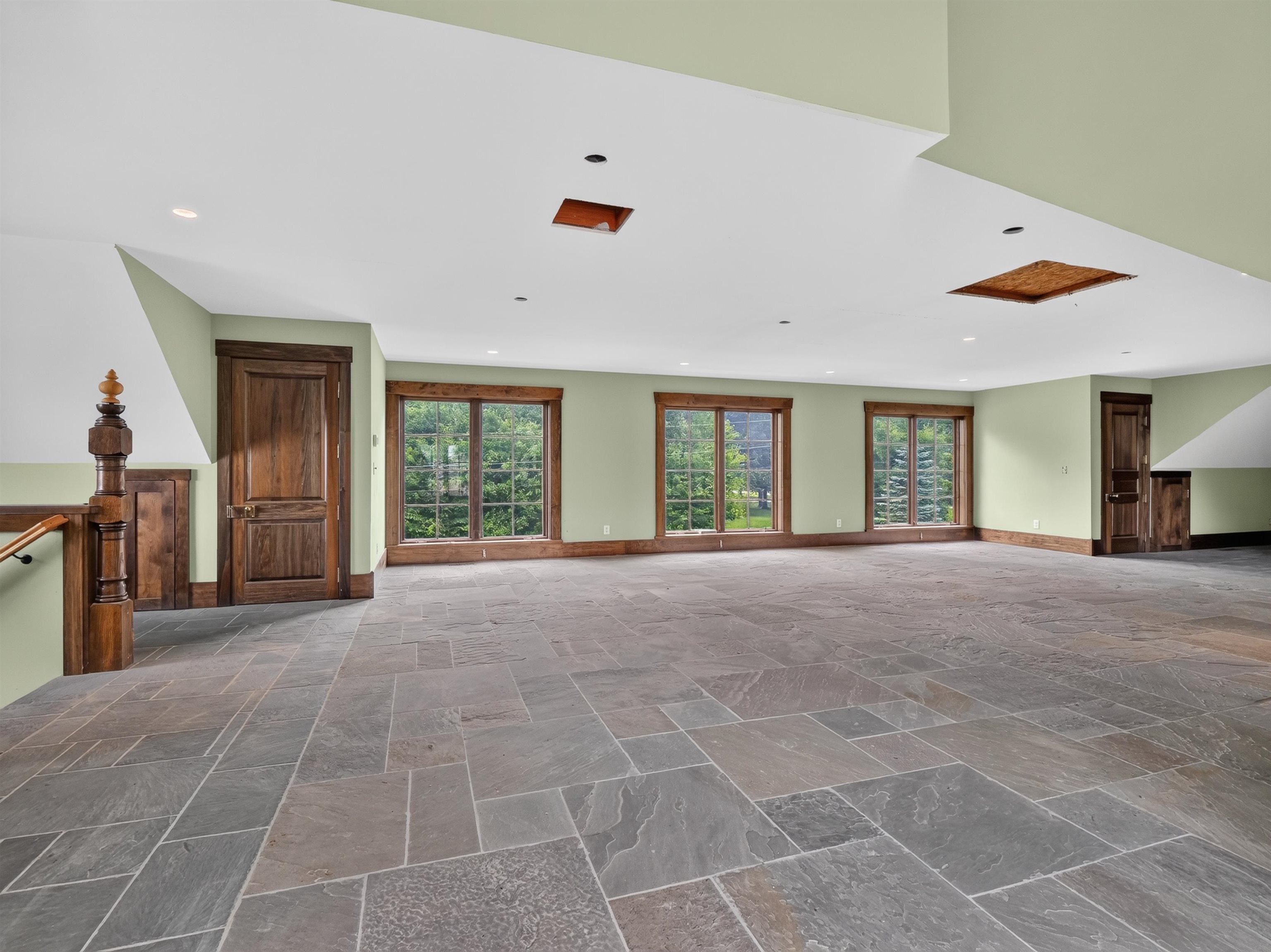 Spacious room featuring natural stone tile flooring, three large windows with wood trim, recessed lighting, and a wood staircase with an ornate newel post