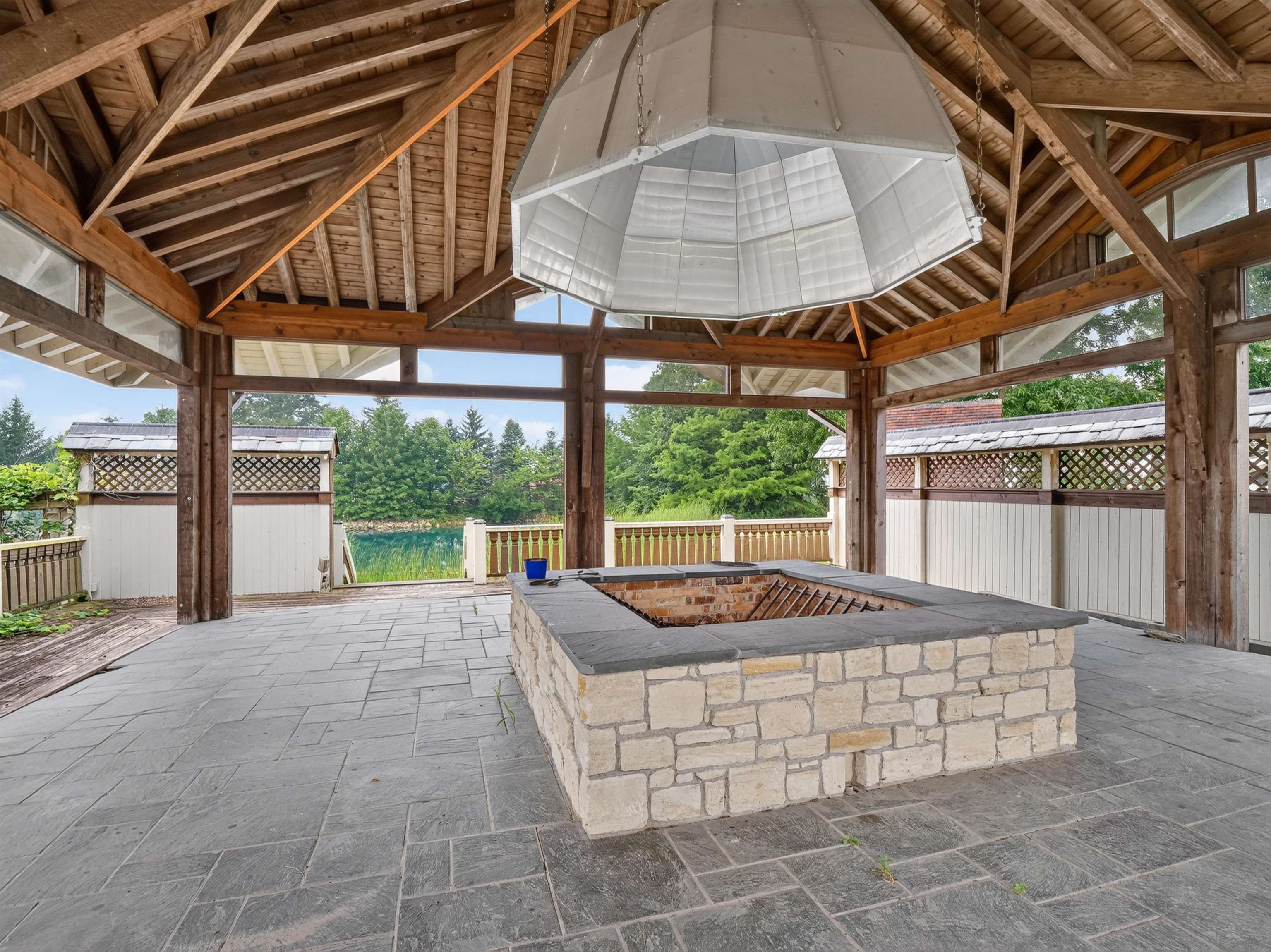 Spacious outdoor pavilion featuring a stone-clad fire pit, exposed timber rafter ceiling, and large format paver flooring