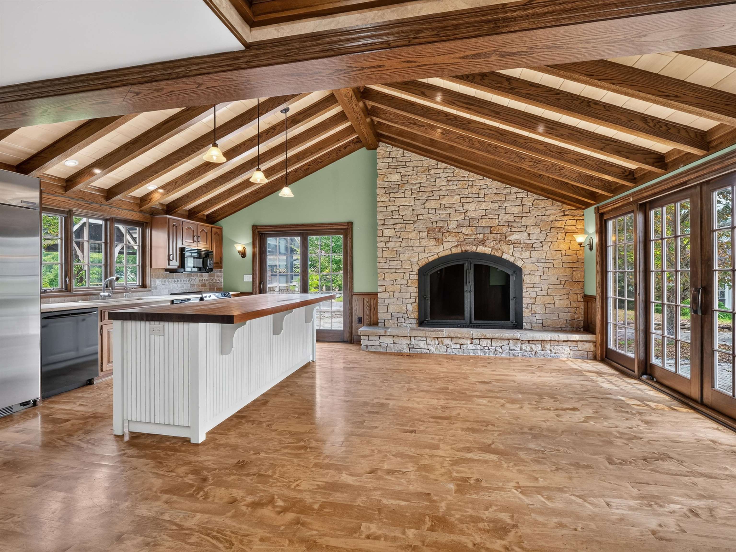 Vaulted wood-beamed ceiling over an open-concept living space featuring a stone fireplace with a black metal insert, a kitchen island with a butcher block countertop, wood-finish flooring, and double French doors