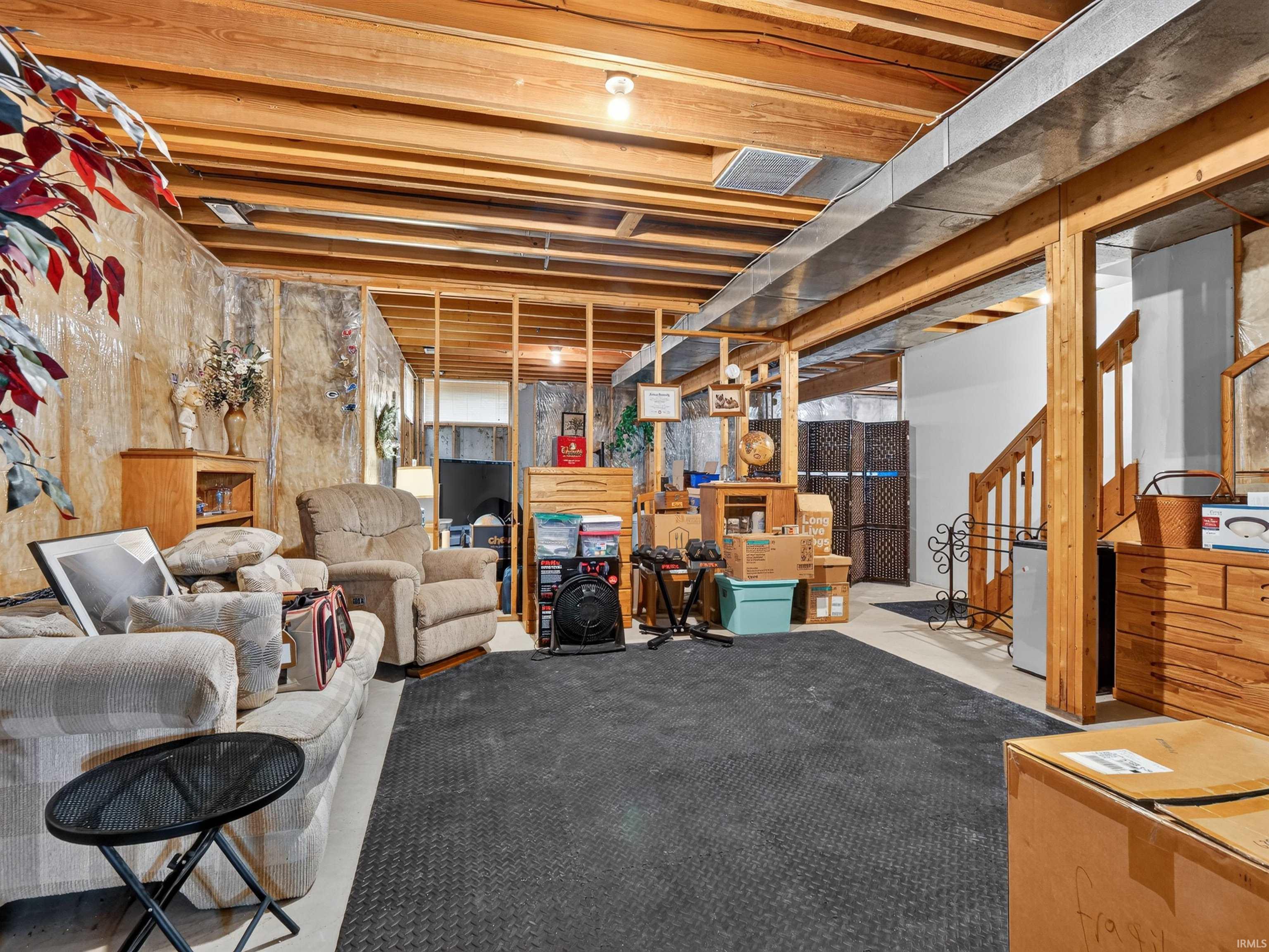 Unfinished basement with exposed wood-frame ceiling and support beams, featuring a partial staircase with wood newel posts and banister, concrete flooring, and exposed insulation on perimeter walls