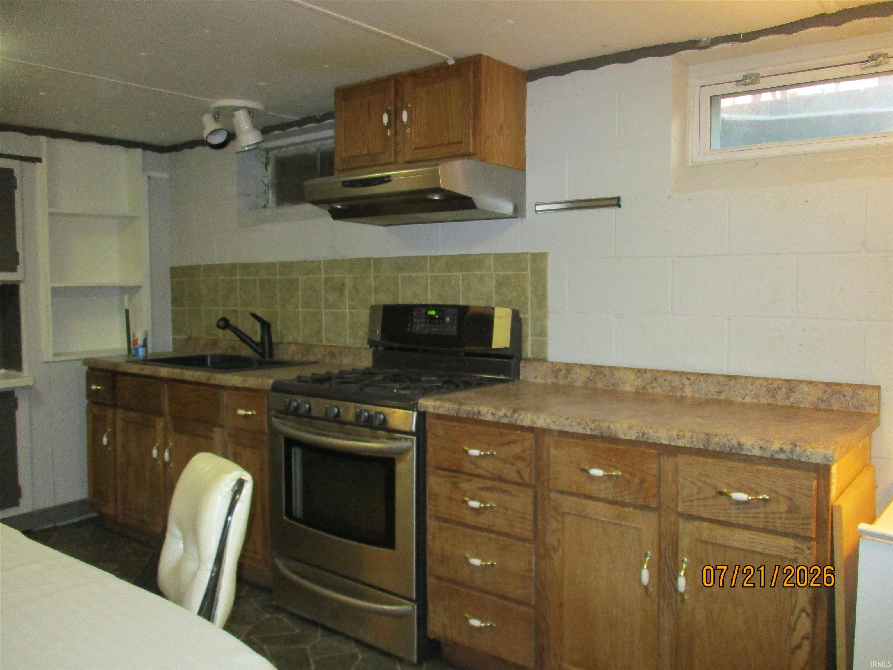 Kitchen area featuring wood-finish cabinetry, stainless steel range and hood, stone-look countertops, and tile backsplash