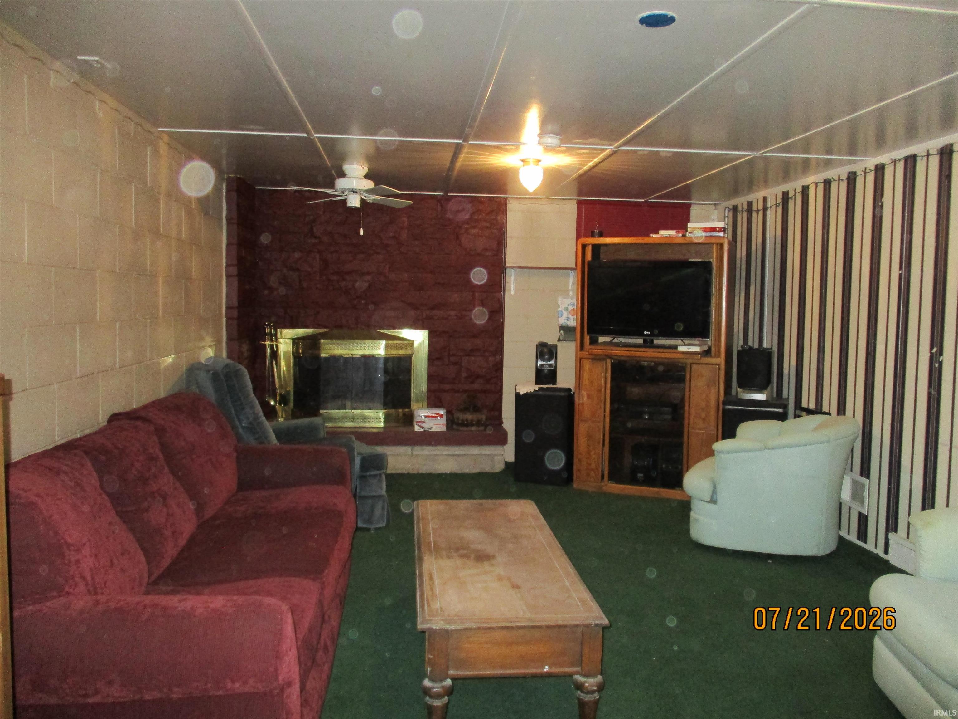 Room featuring a stone fireplace with a brass firebox, green carpet flooring, a drop ceiling with integrated lighting, and a ceiling fan