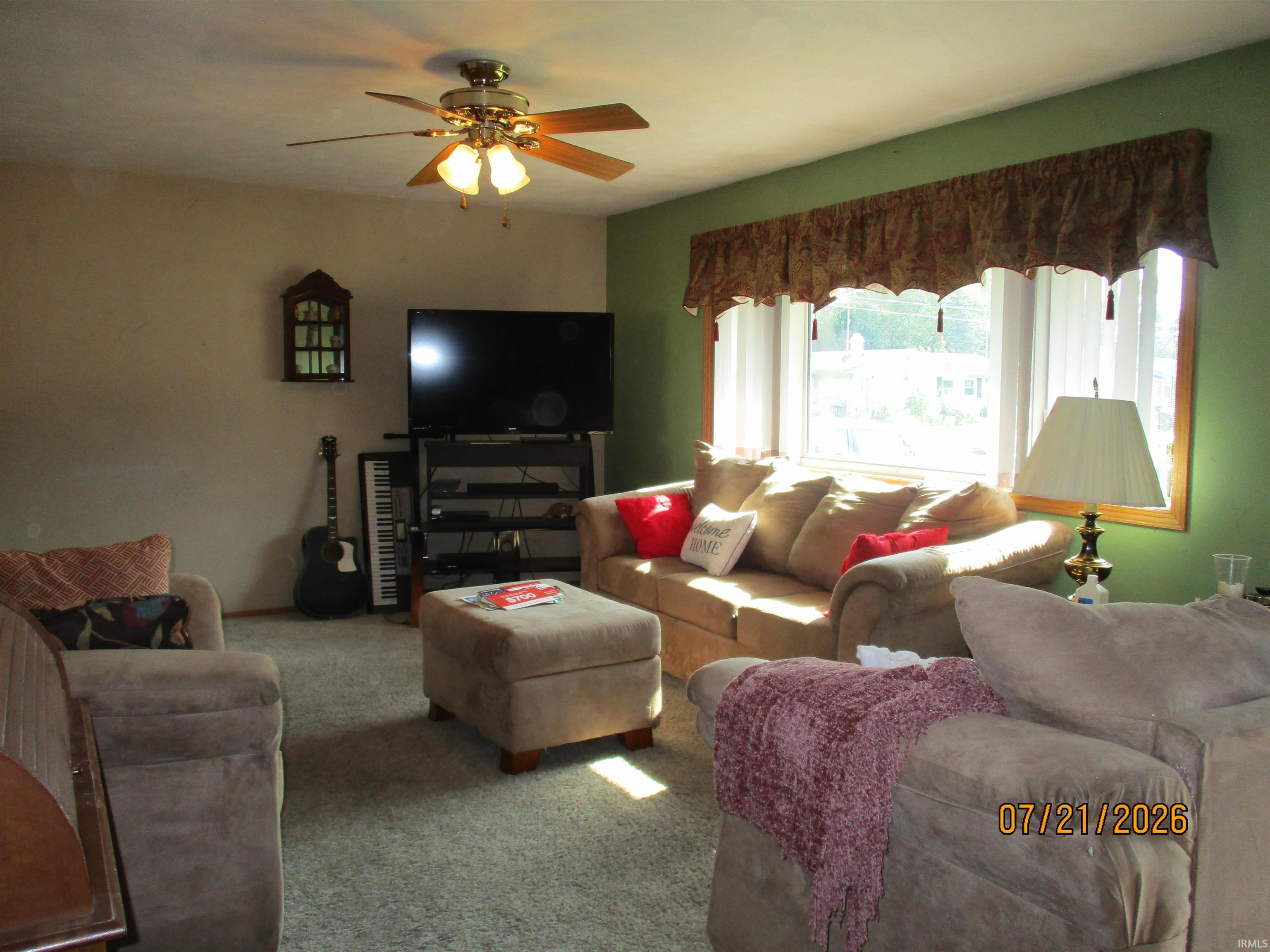 Living area featuring neutral-toned carpeting, a multi-pane window with wood-frame trim, and a ceiling fan with integrated lighting