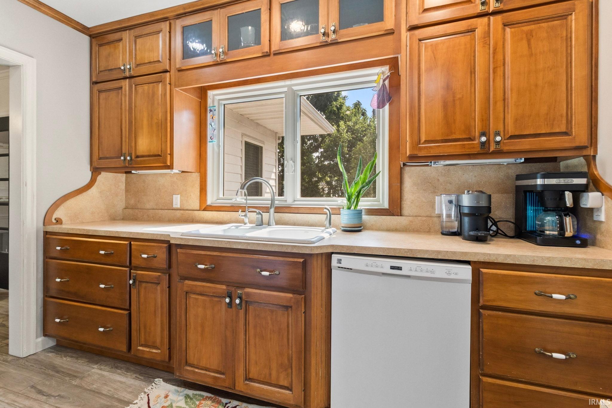 Kitchen featuring wood cabinetry, a double basin sink with a gooseneck faucet, light-toned countertops, and a built-in dishwasher