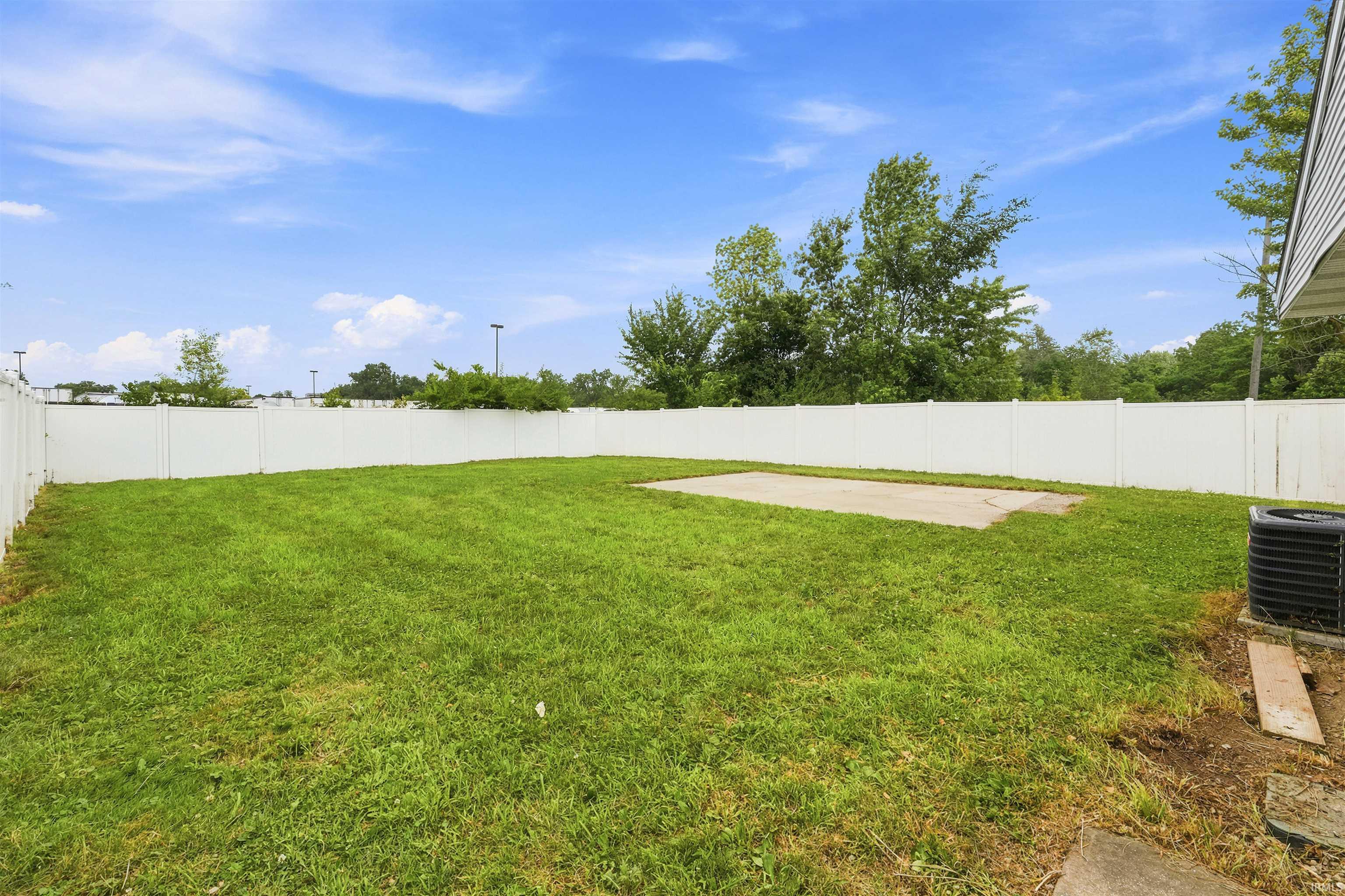 Expansive grassy yard enclosed by a white privacy fence, featuring a concrete pad and mature perimeter trees