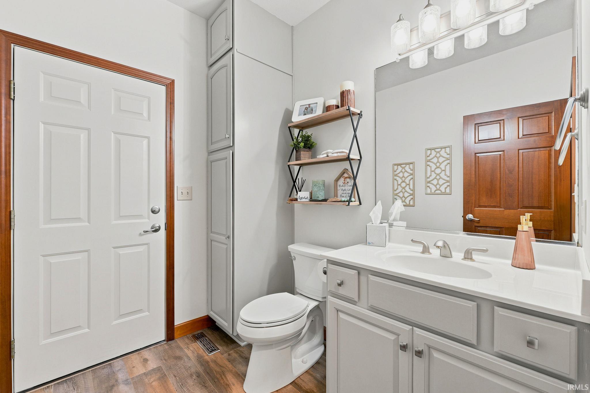 Bathroom featuring wood-finish flooring, a white paneled door with wood trim, a built-in floor-to-ceiling cabinet, a toilet, a vanity with a white countertop and undermount sink, and a large wall mirror
