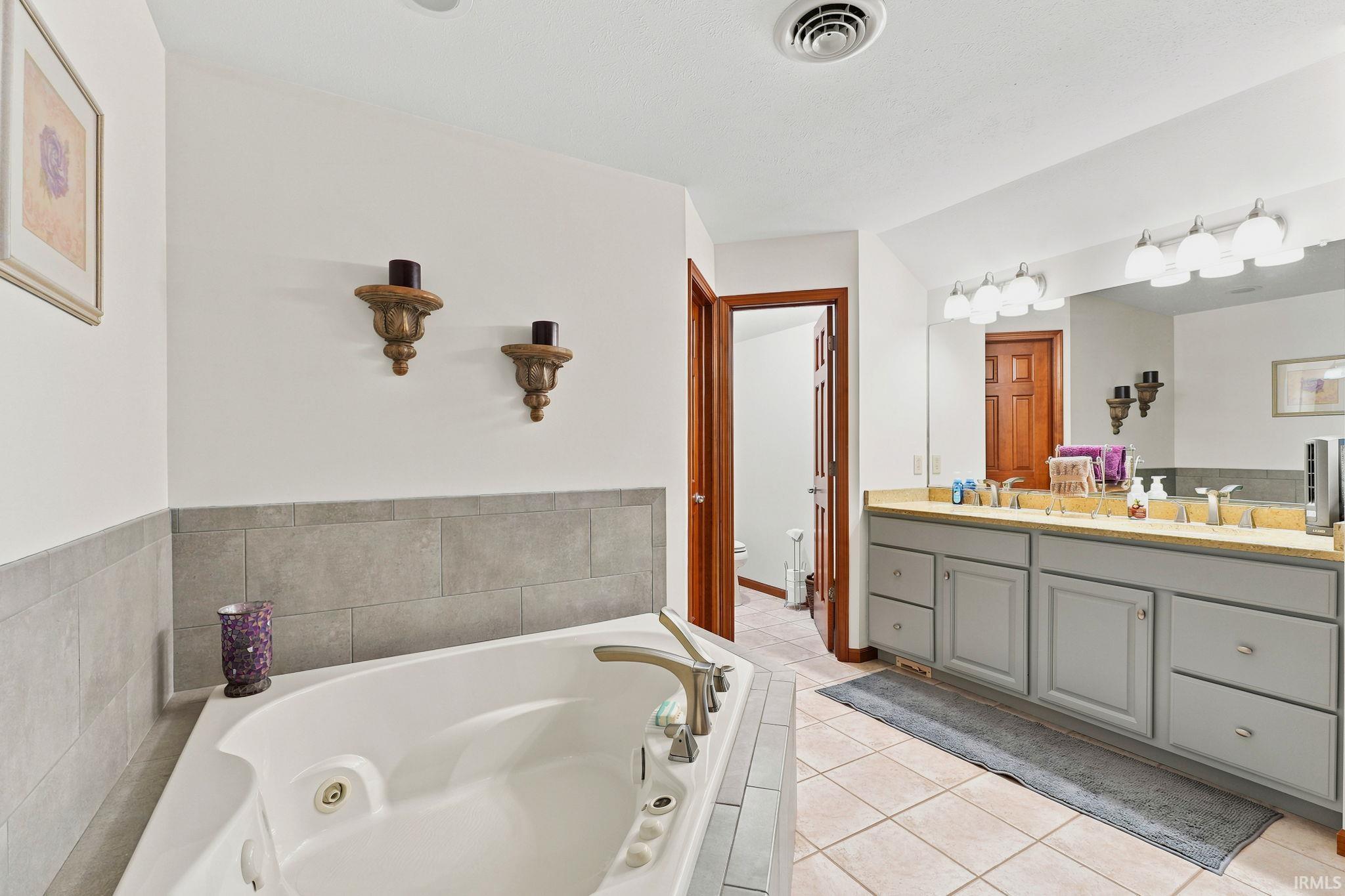 Bathroom featuring a built-in corner soaking tub with deck-mounted fixtures, a dual vanity with light gray cabinetry and a light-toned countertop, and tile flooring
