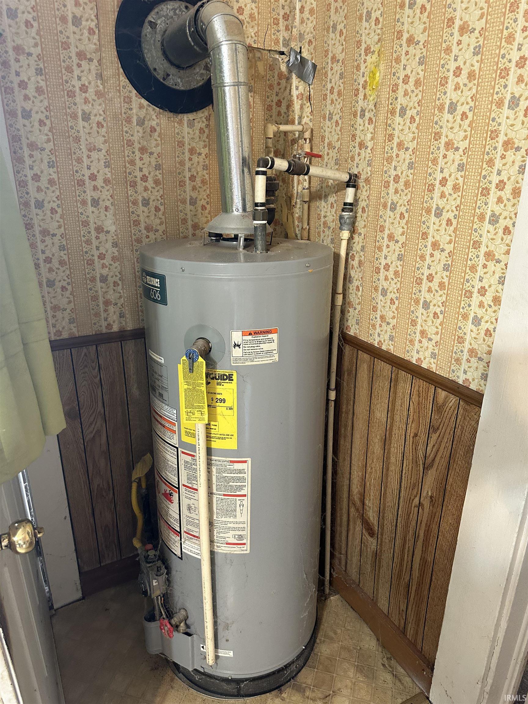 Utility closet featuring a water heater with visible piping and exhaust vent, wood-finish paneling, and patterned wallpaper