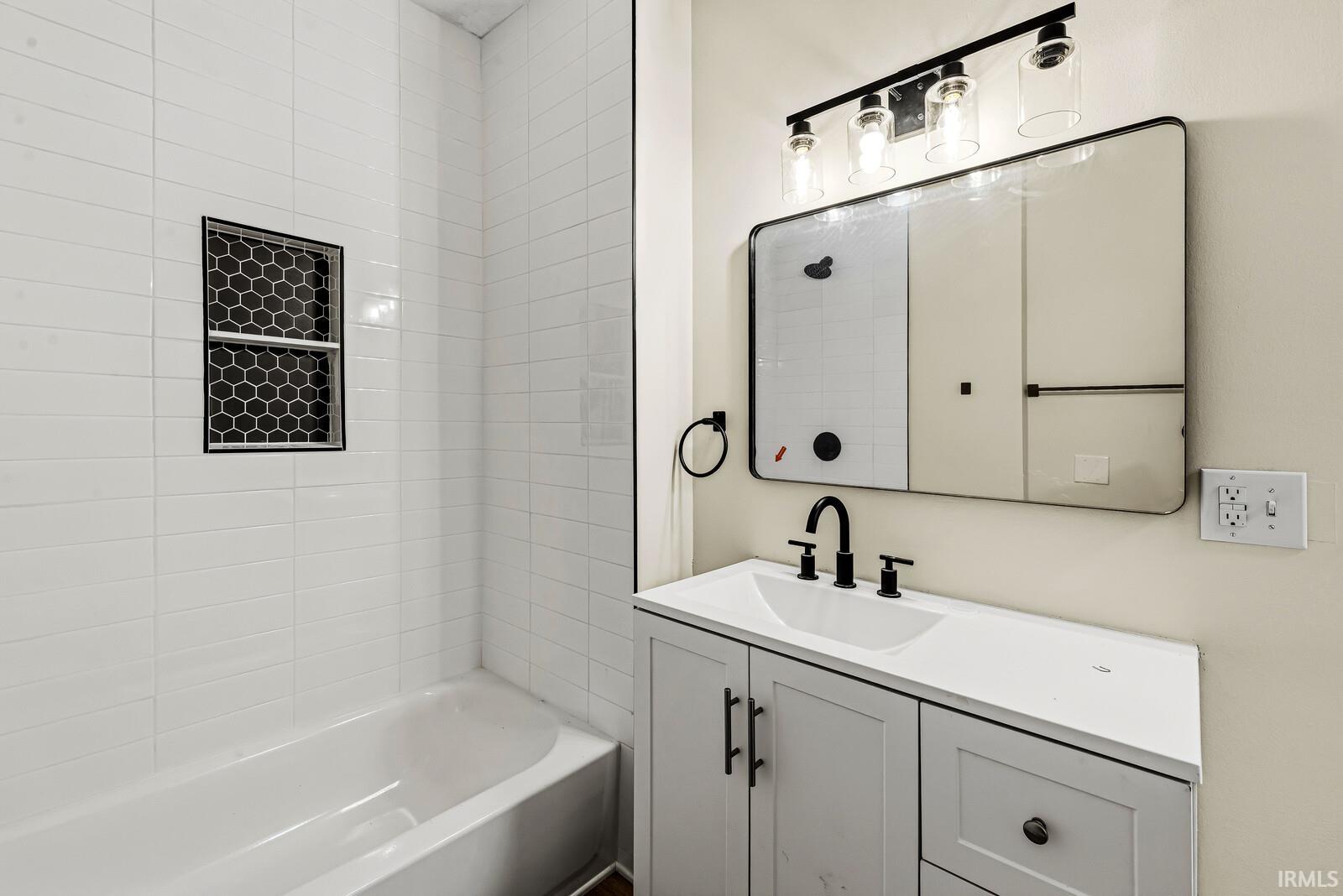 Bathroom featuring a white subway tile tub surround, a recessed shower niche with dark hexagonal tile, a single vanity with a white countertop, a rectangular sink with matte black fixtures, and a framed mirror with integrated lighting