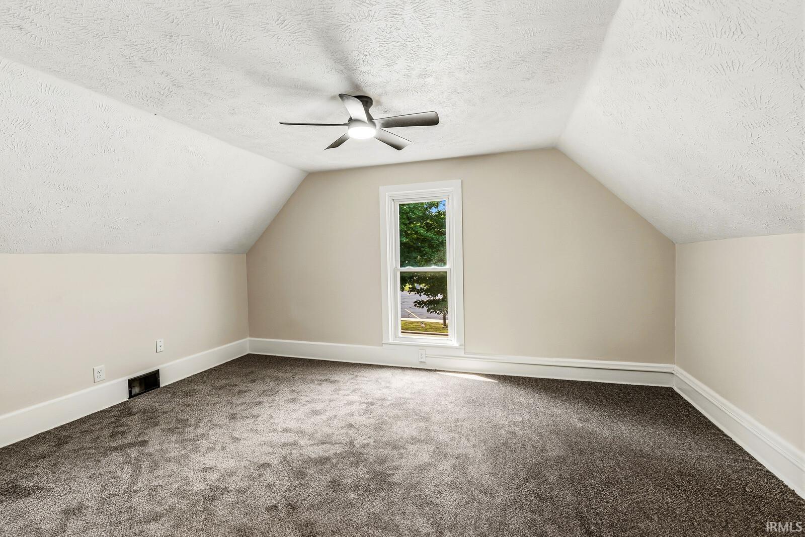 Attic space featuring neutral wall paint, textured ceiling, a ceiling fan with integrated lighting, window with white trim, and gray carpet flooring