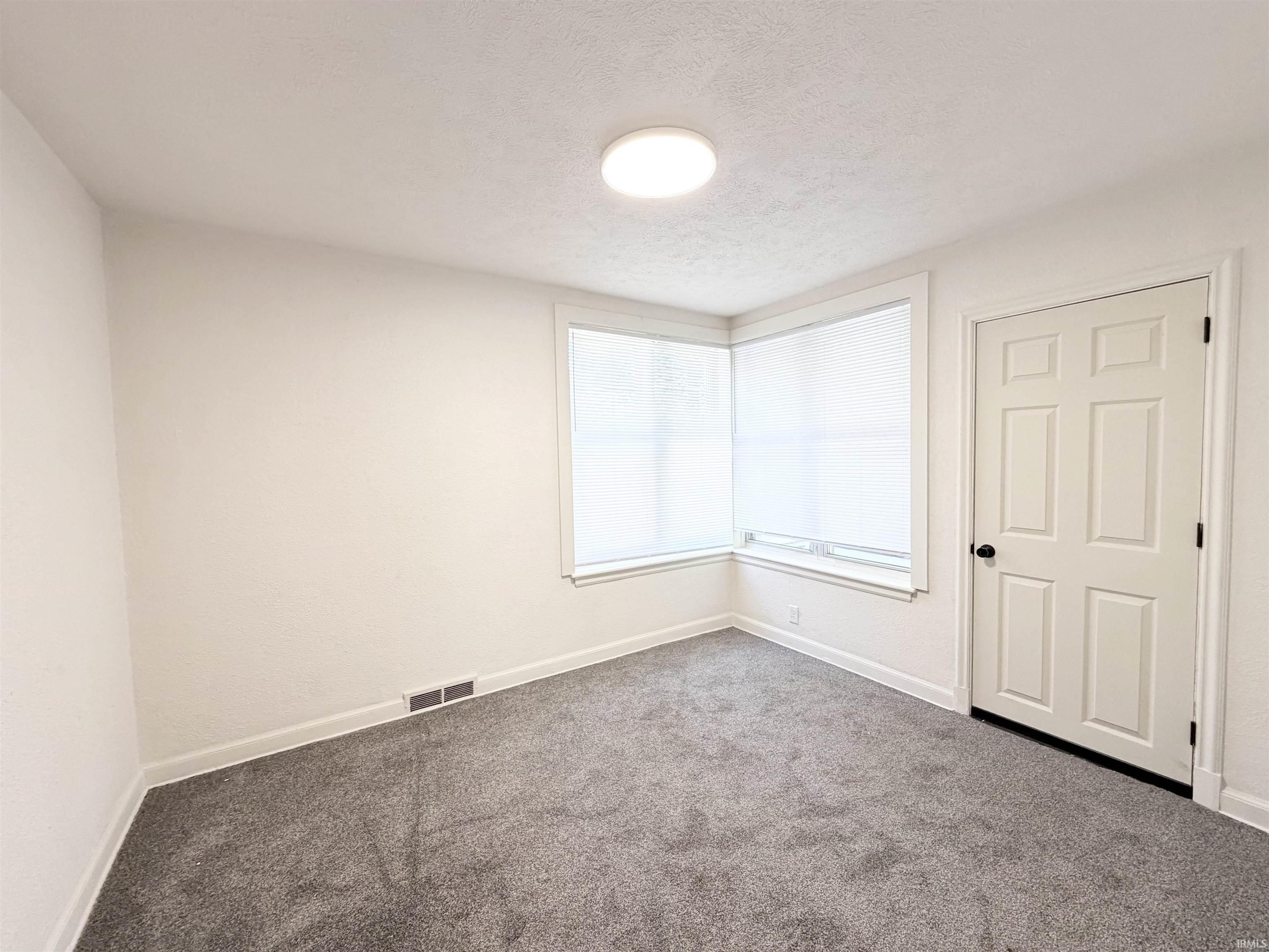 Room featuring corner windows with white roller blinds, gray carpet, a white paneled door with black hardware, and a ceiling-mounted flush light fixture