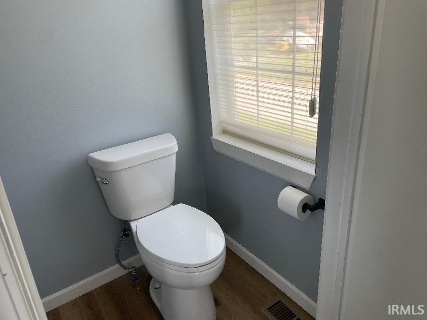 White toilet with chrome flush lever, window with blinds, wood-finish flooring, and light blue walls