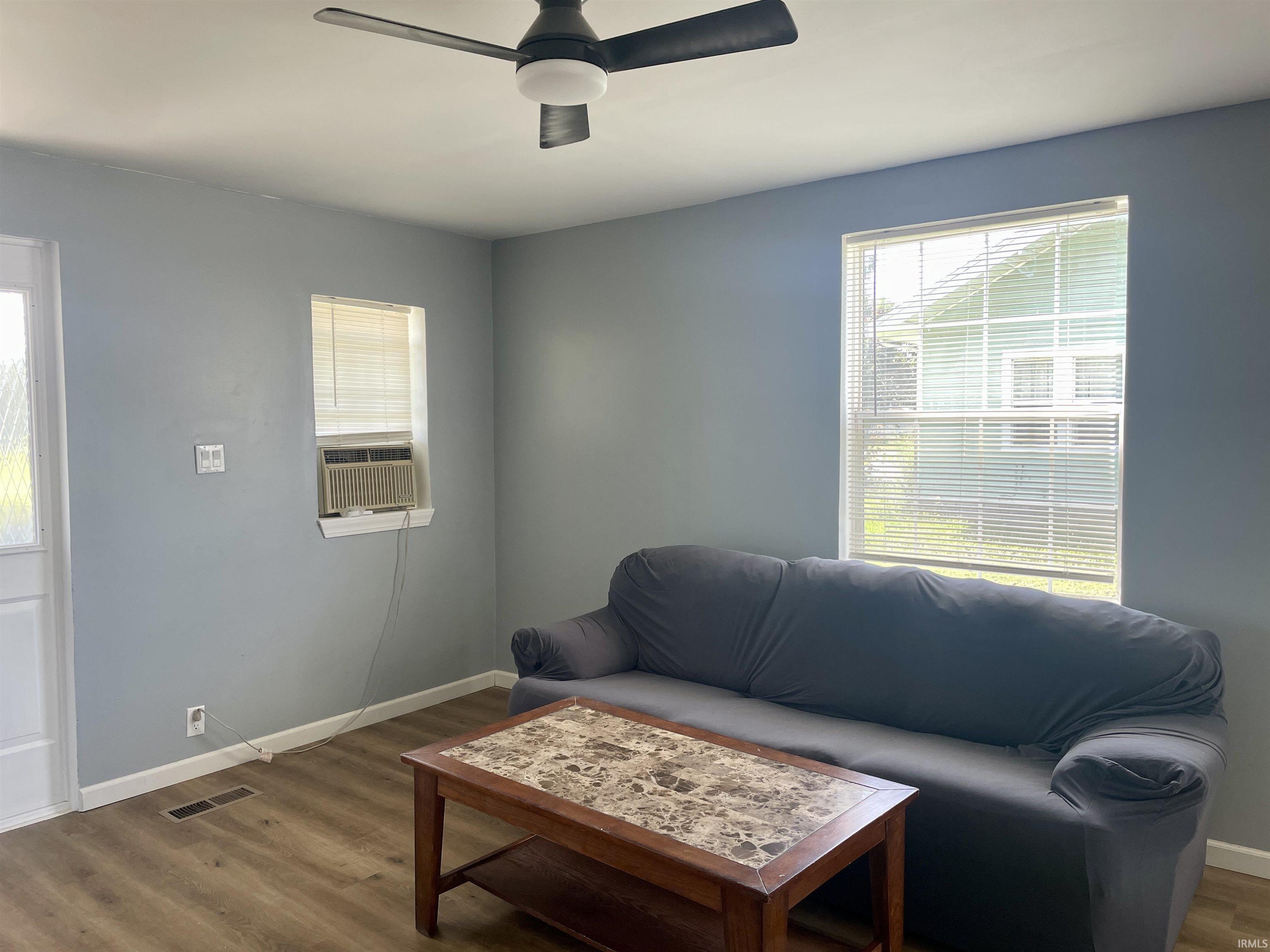 Living area with wood-finish flooring, two windows with blinds, a ceiling fan, and painted walls