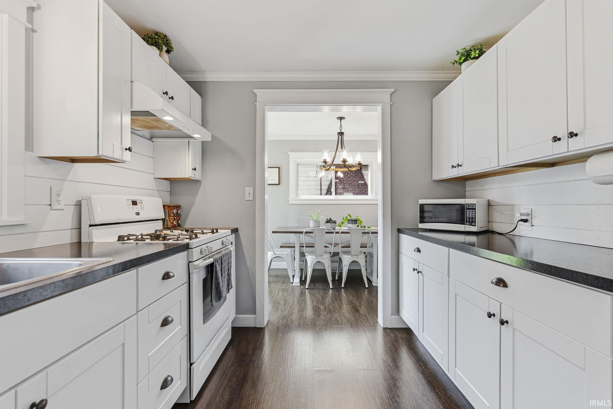 Galley kitchen featuring white cabinetry, dark countertops, wood-finish flooring, white shiplap backsplash, and a pass-through opening to a dining area