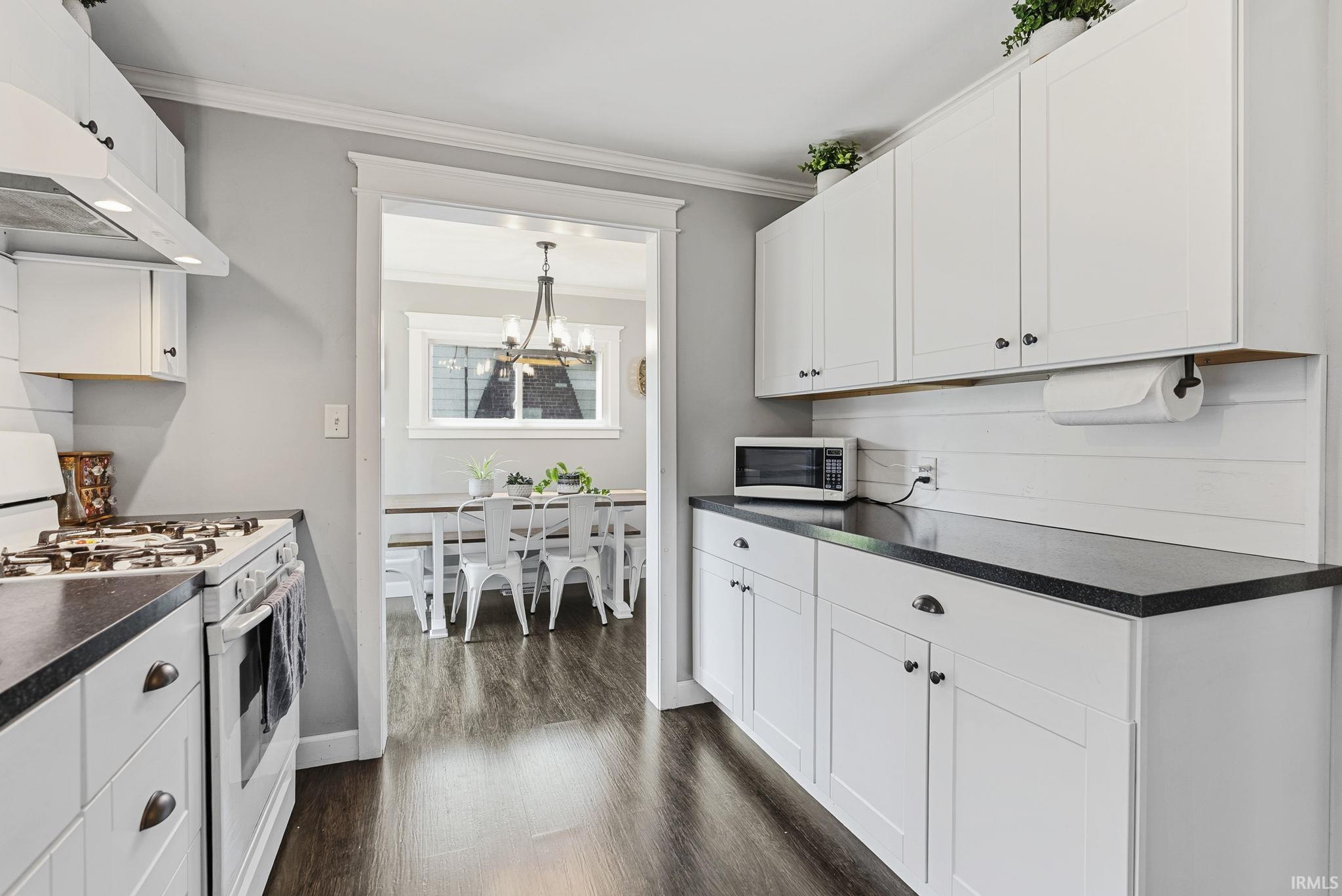 Kitchen featuring white shaker-style cabinetry, dark countertops, white shiplap-style backsplash, wood-finish flooring, and a gas range with an overhead vent hood