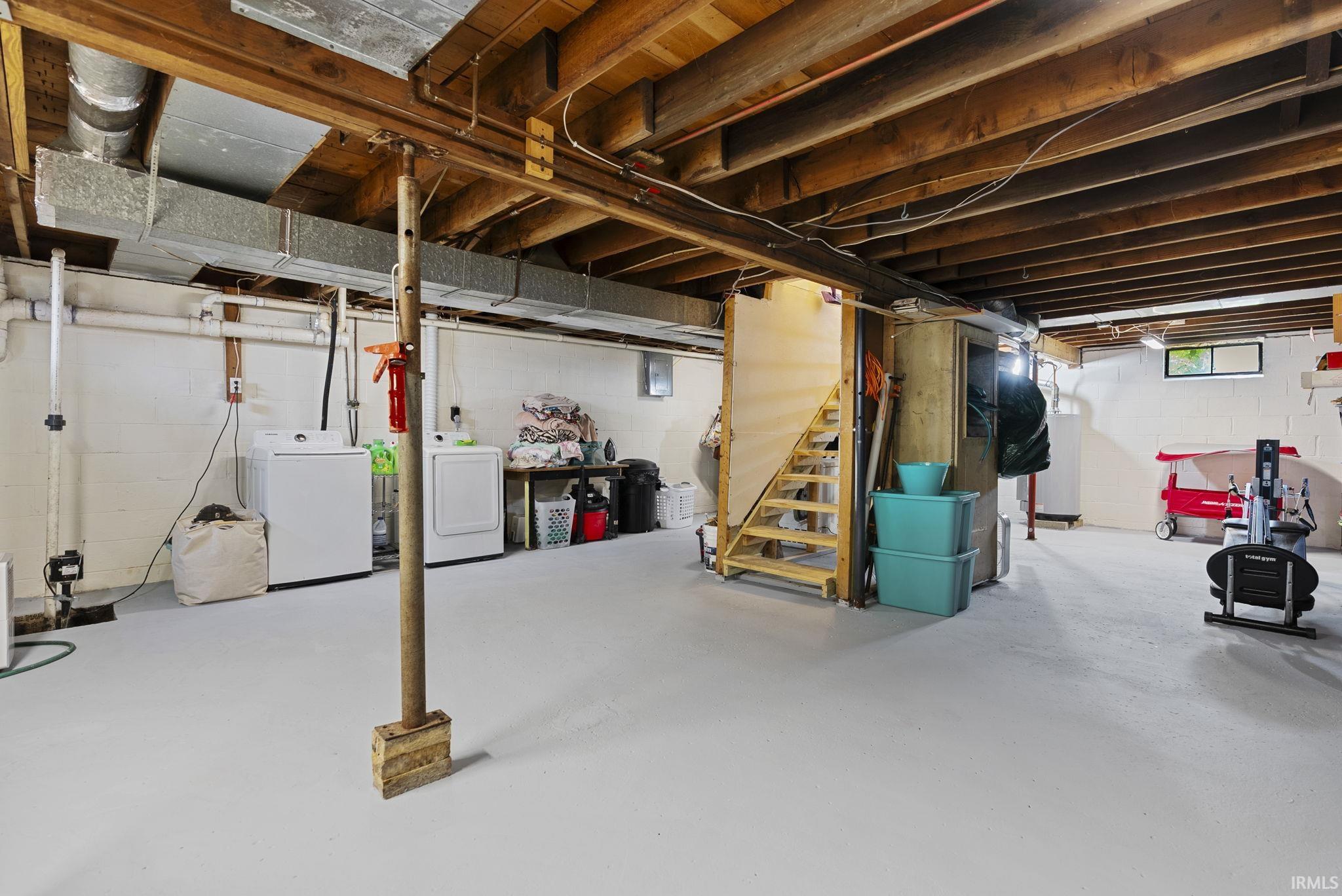 Spacious utility room featuring a painted concrete floor, exposed wood ceiling beams, white block walls, and dedicated laundry area