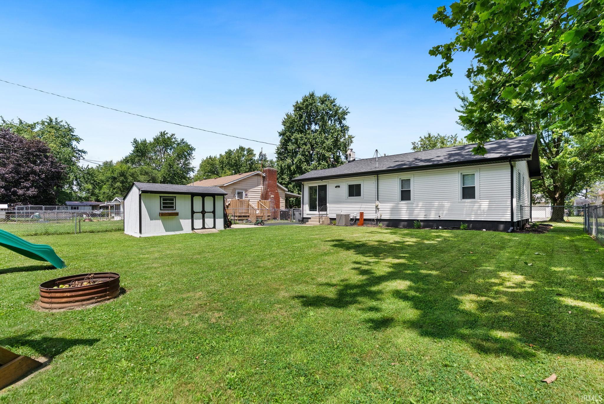 Expansive backyard featuring a manicured lawn, a white storage shed with black trim, mature trees, and a chain-link fence bordering the property
